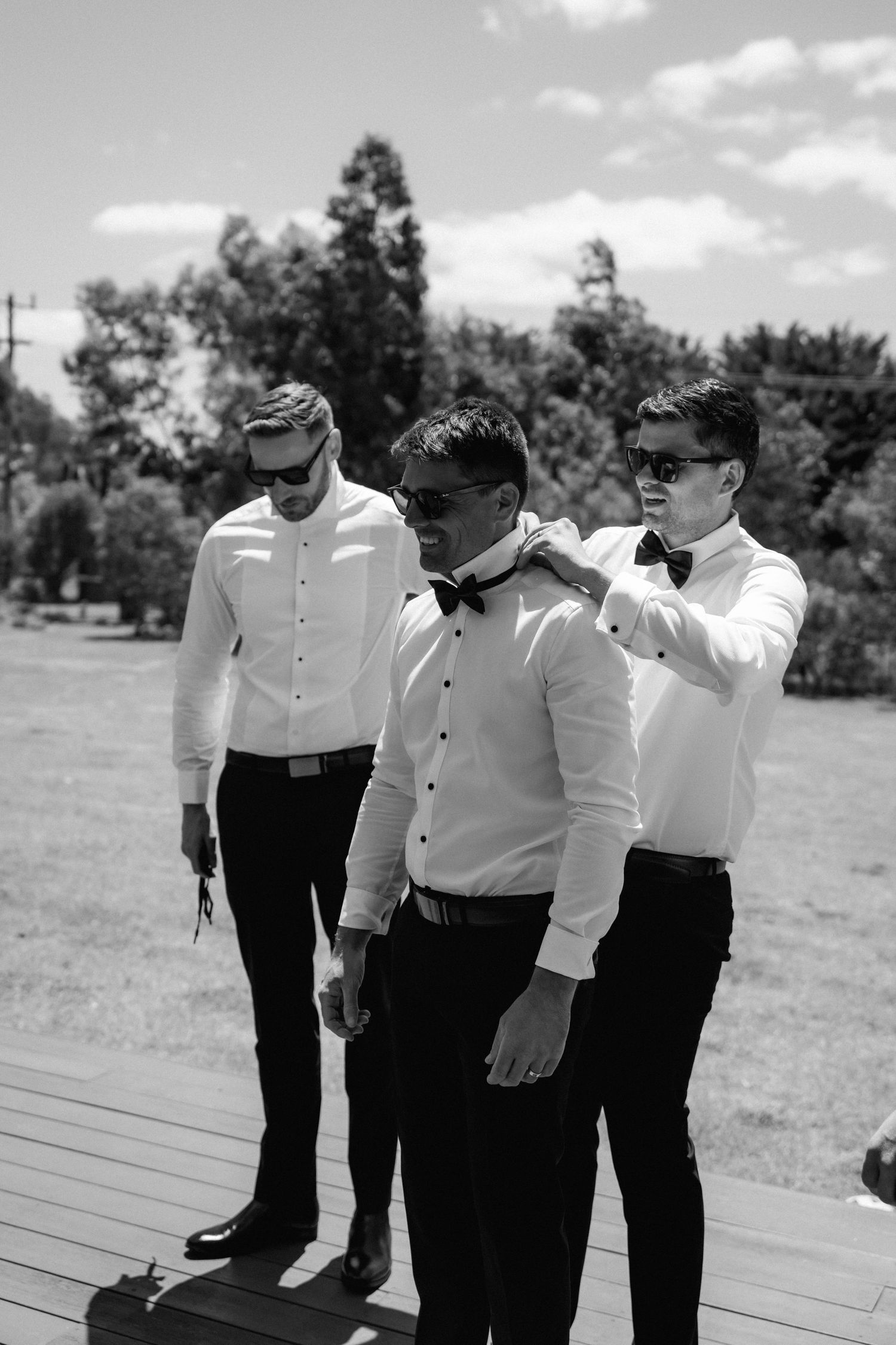 Groomsmen in matching white shirts and black pants share a candid moment outdoors.
