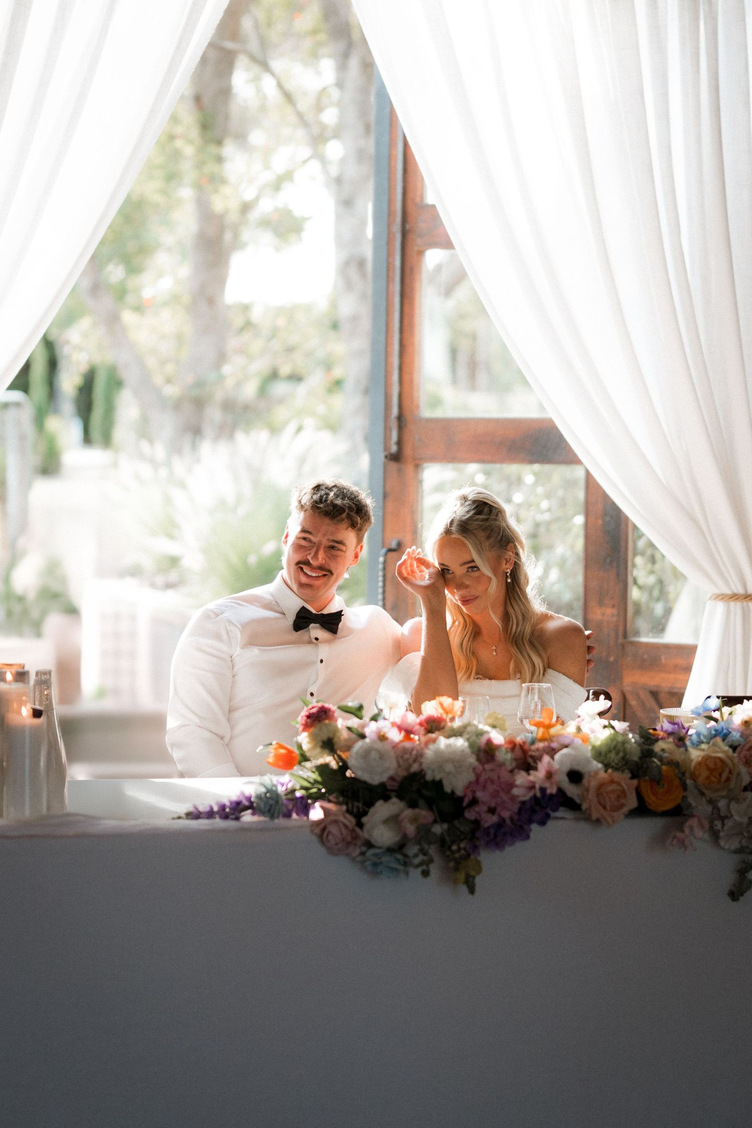 Bride and groom laughing at sweetheart table adorned with colorful floral arrangements.