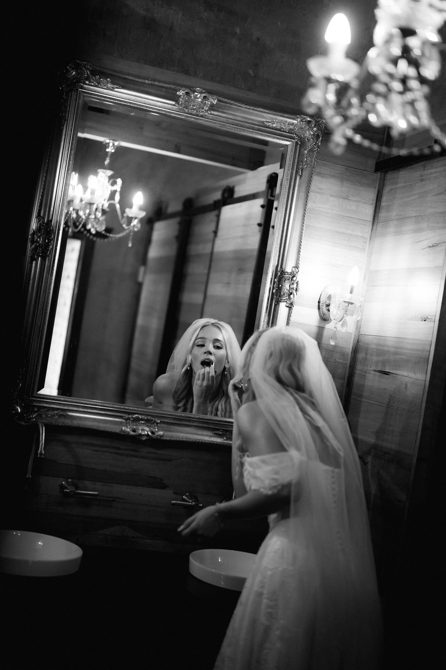 Black and white photo of blonde bride applying makeup at ornate mirror.
