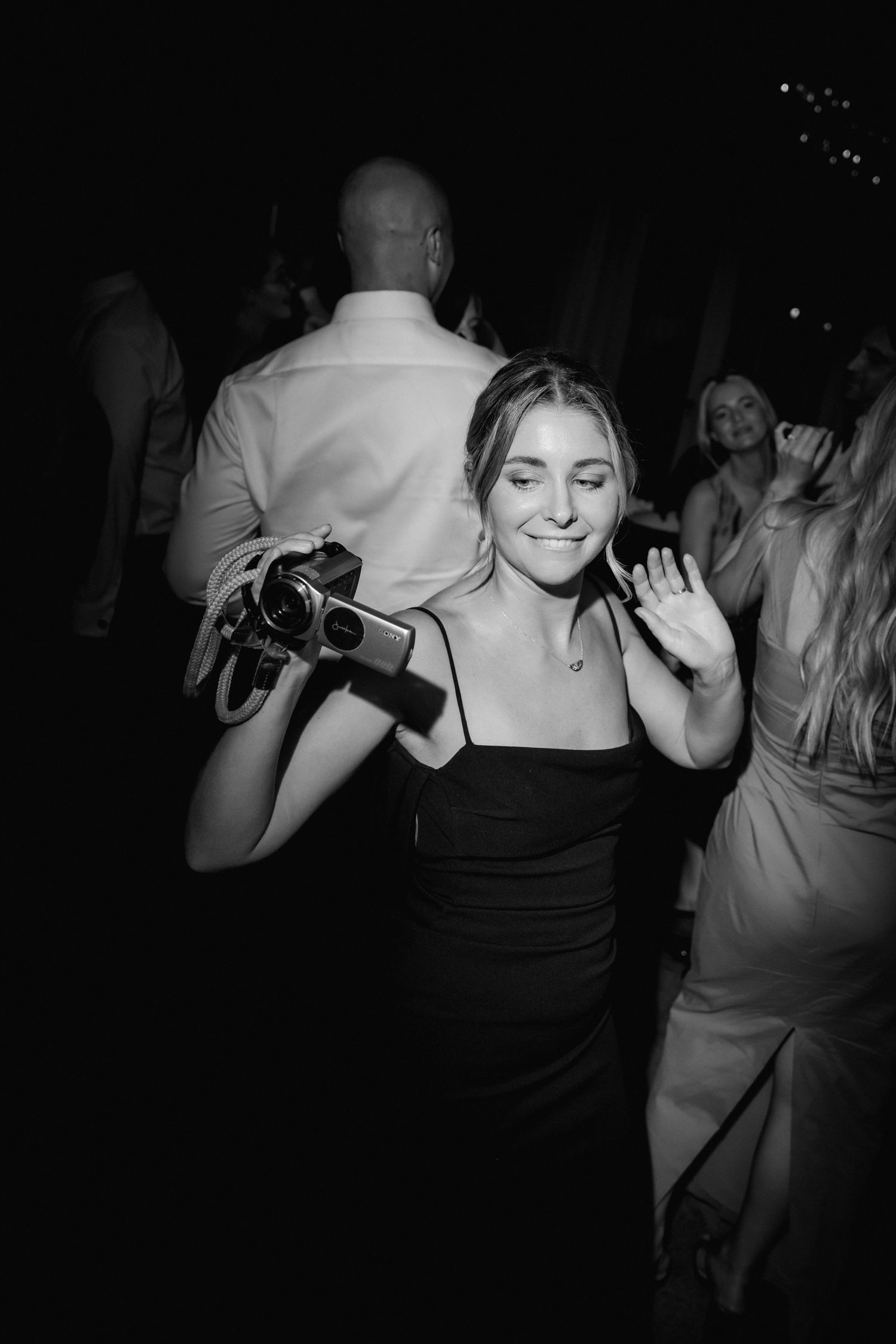 Black and white photo of someone in a black dress holding a camera and waving at a party.