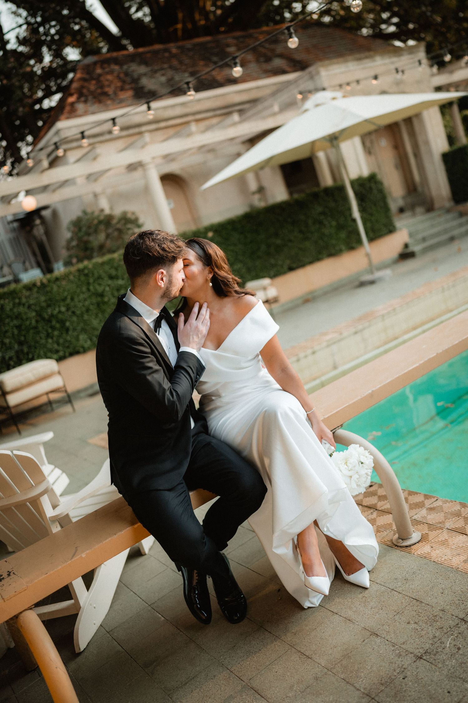 Romantic couple sits poolside at an outdoor venue with string lights overhead and white umbrellas in the background.