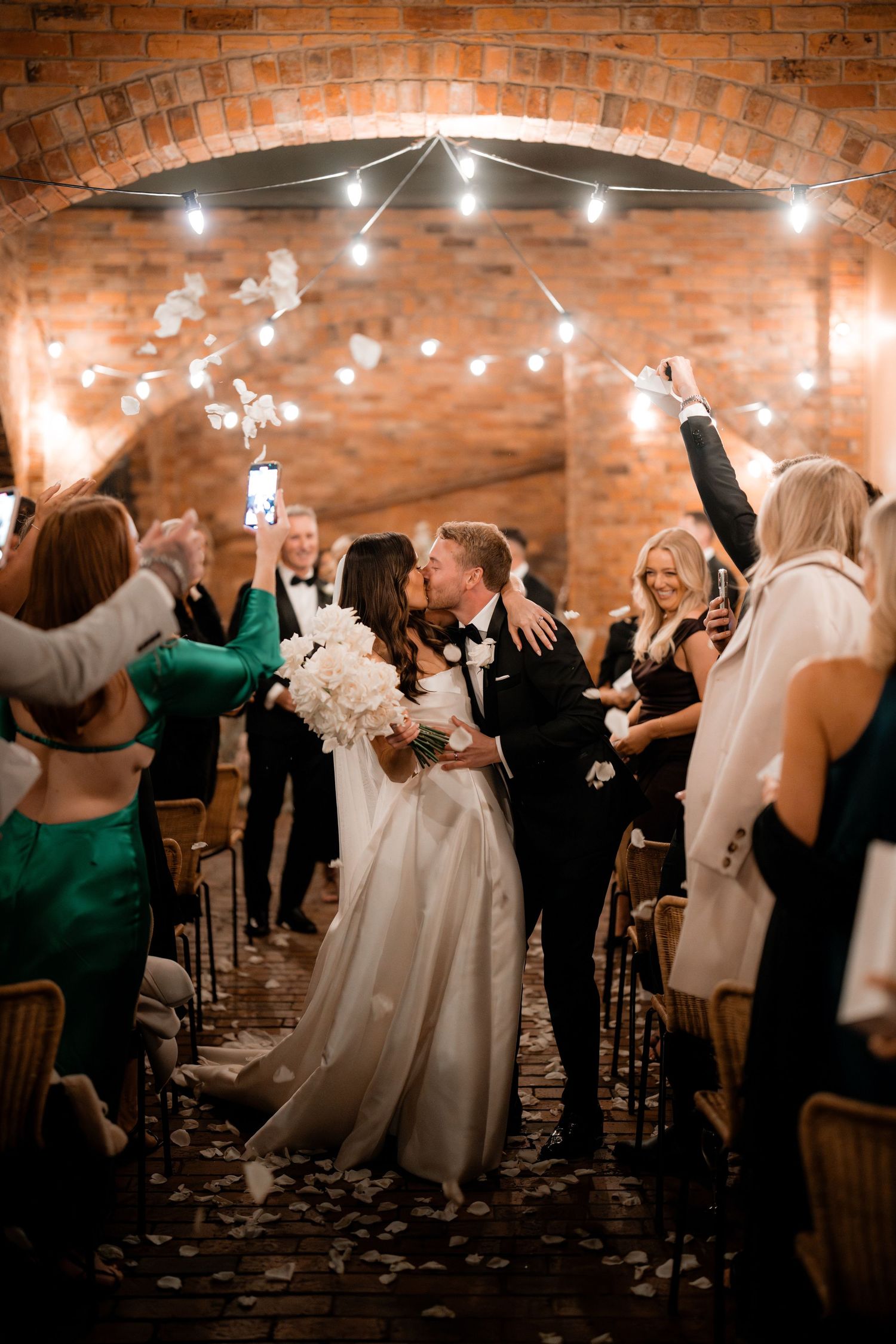 Newlywed couple shares romantic kiss under brick archway as guests celebrate with sparklers at wedding reception.