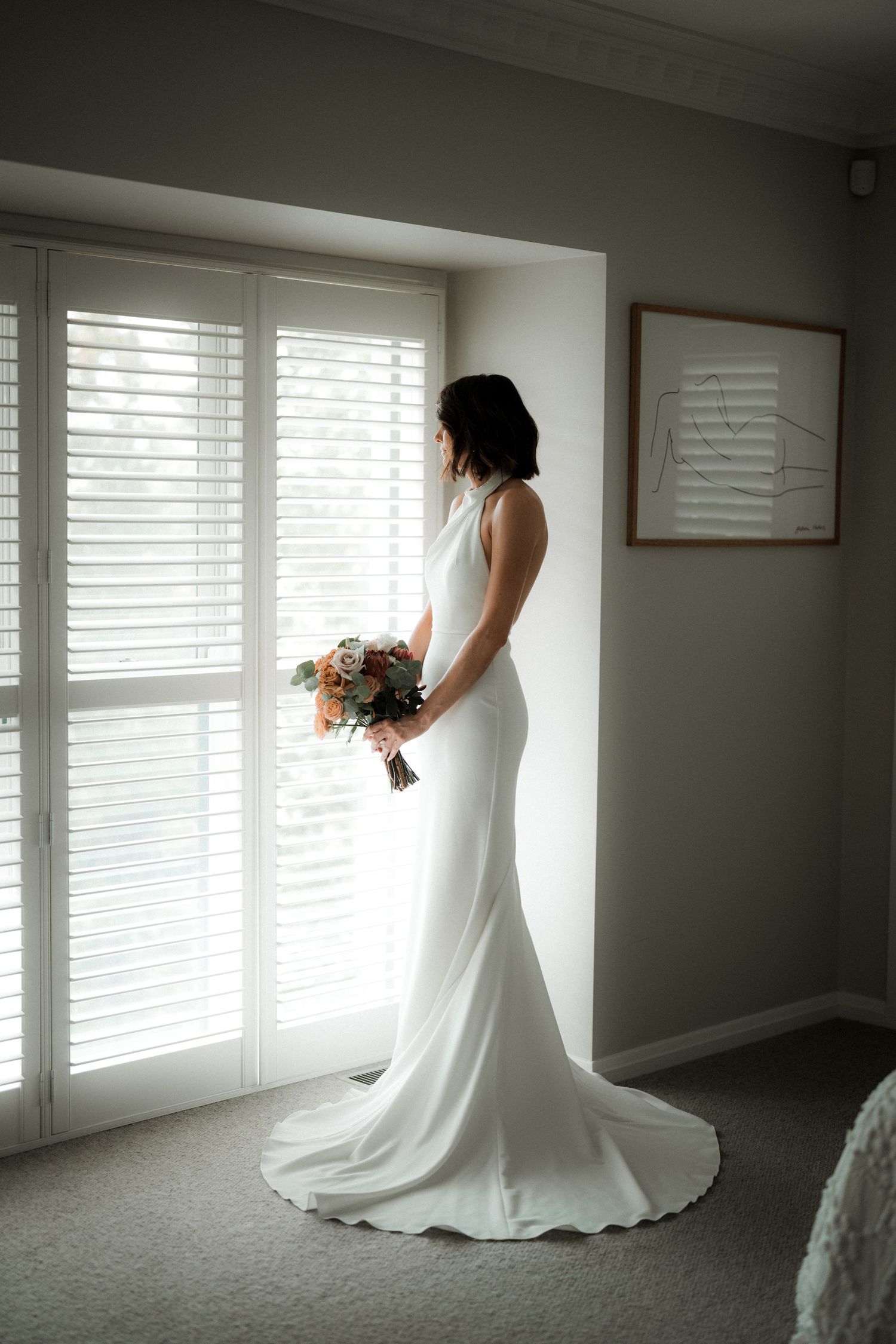 Classic elegant white wedding dress silhouetted against bright window with cascading bouquet in modern interior.