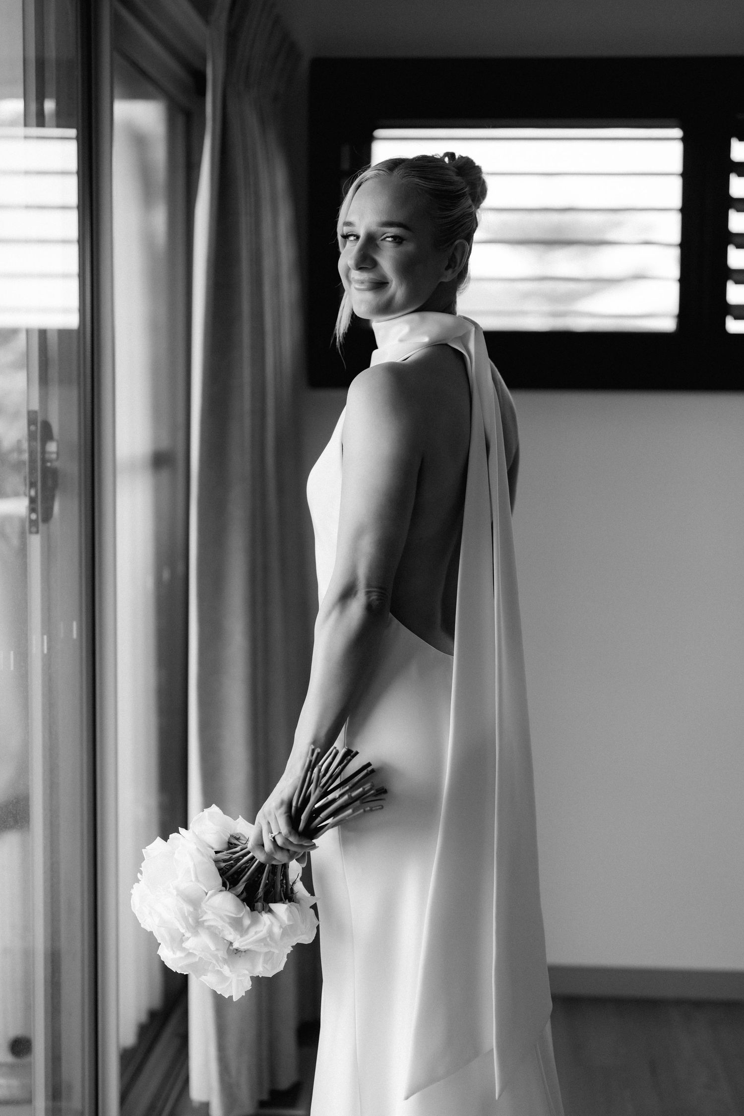 Black and white photo of bride in sleek wedding dress with open back holding white bouquet by window.