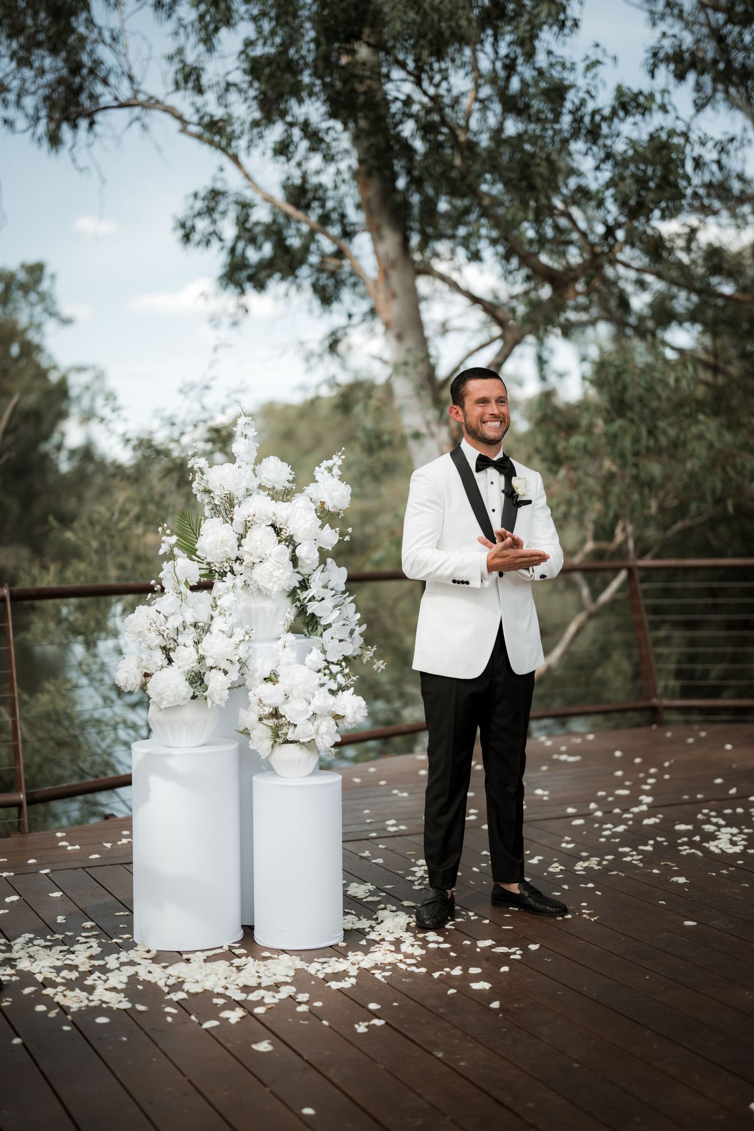 White floral arrangements in vases on a wooden deck with scattered petals.