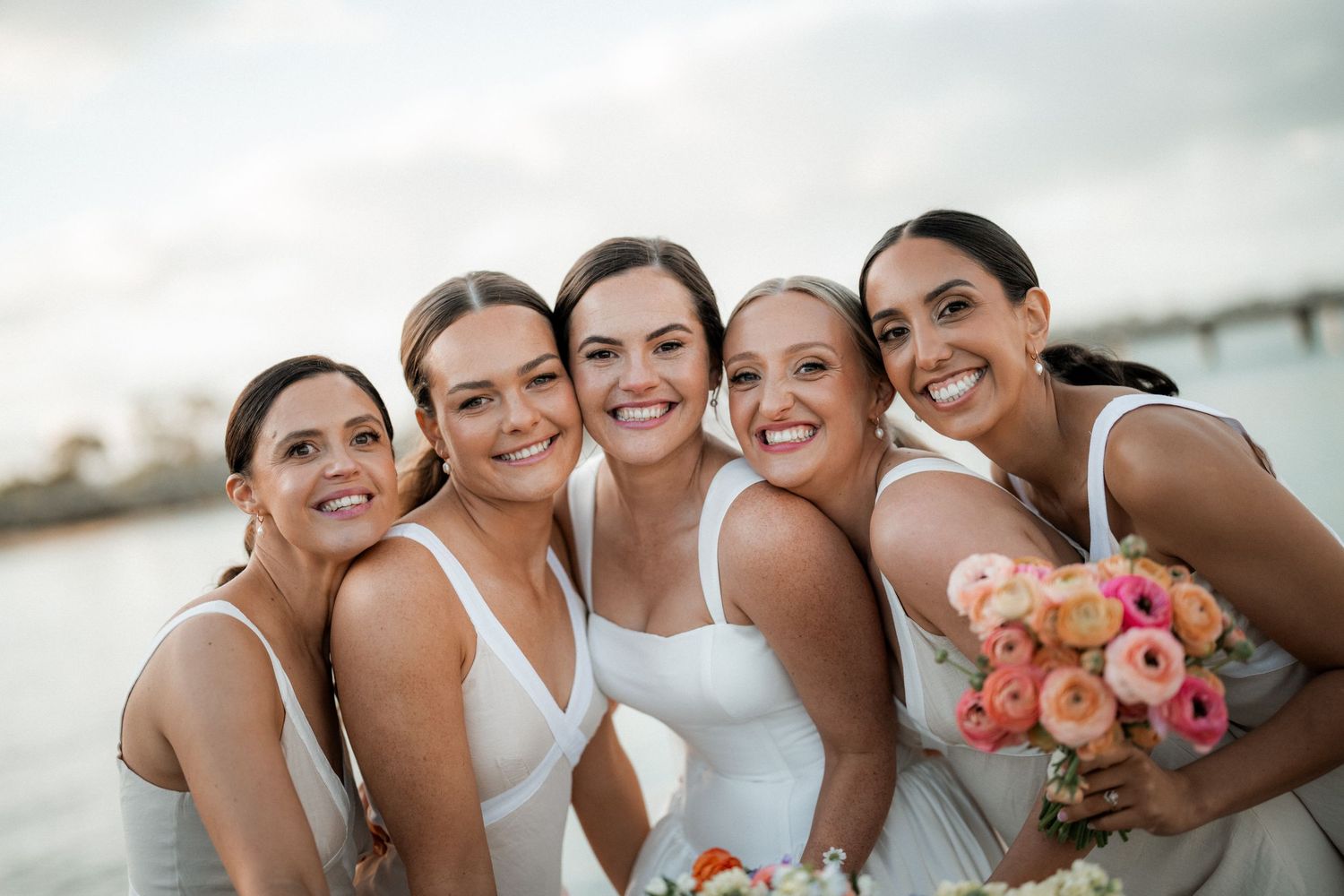 A group of bridesmaids in matching dresses pose together on a beach holding pink floral bouquets.