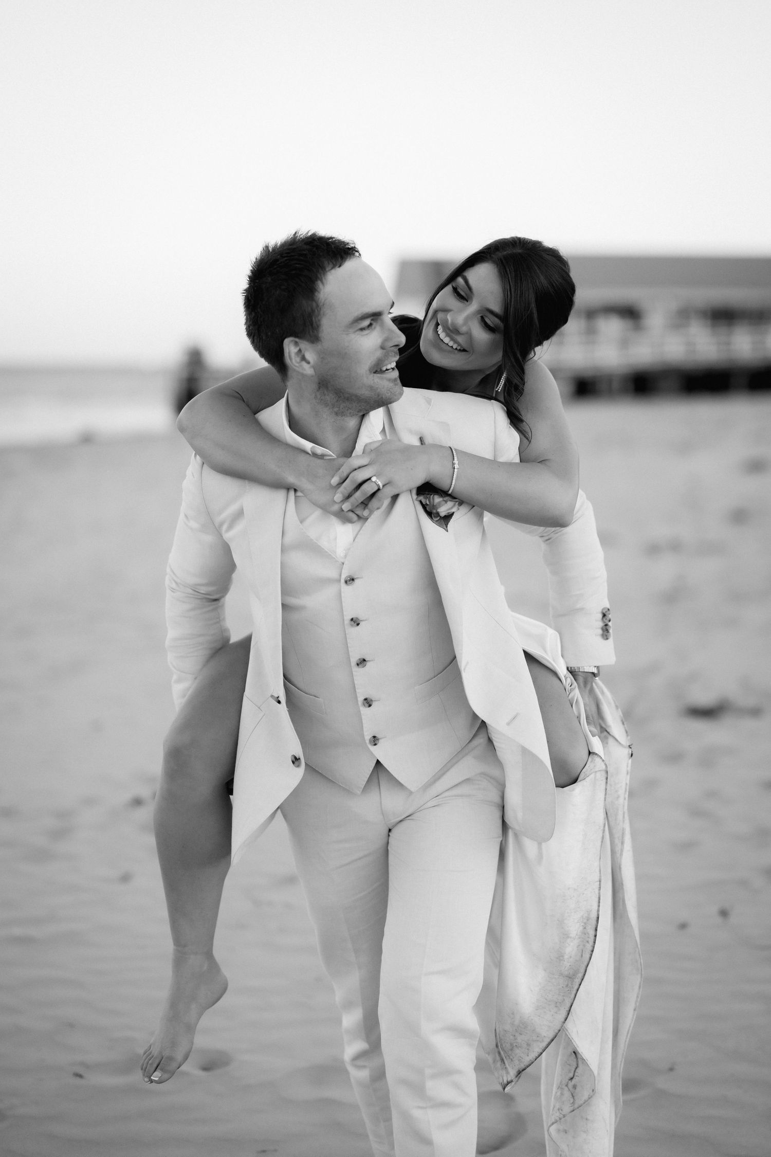 A couple in white formal wear embraces on a beach during a romantic photoshoot.