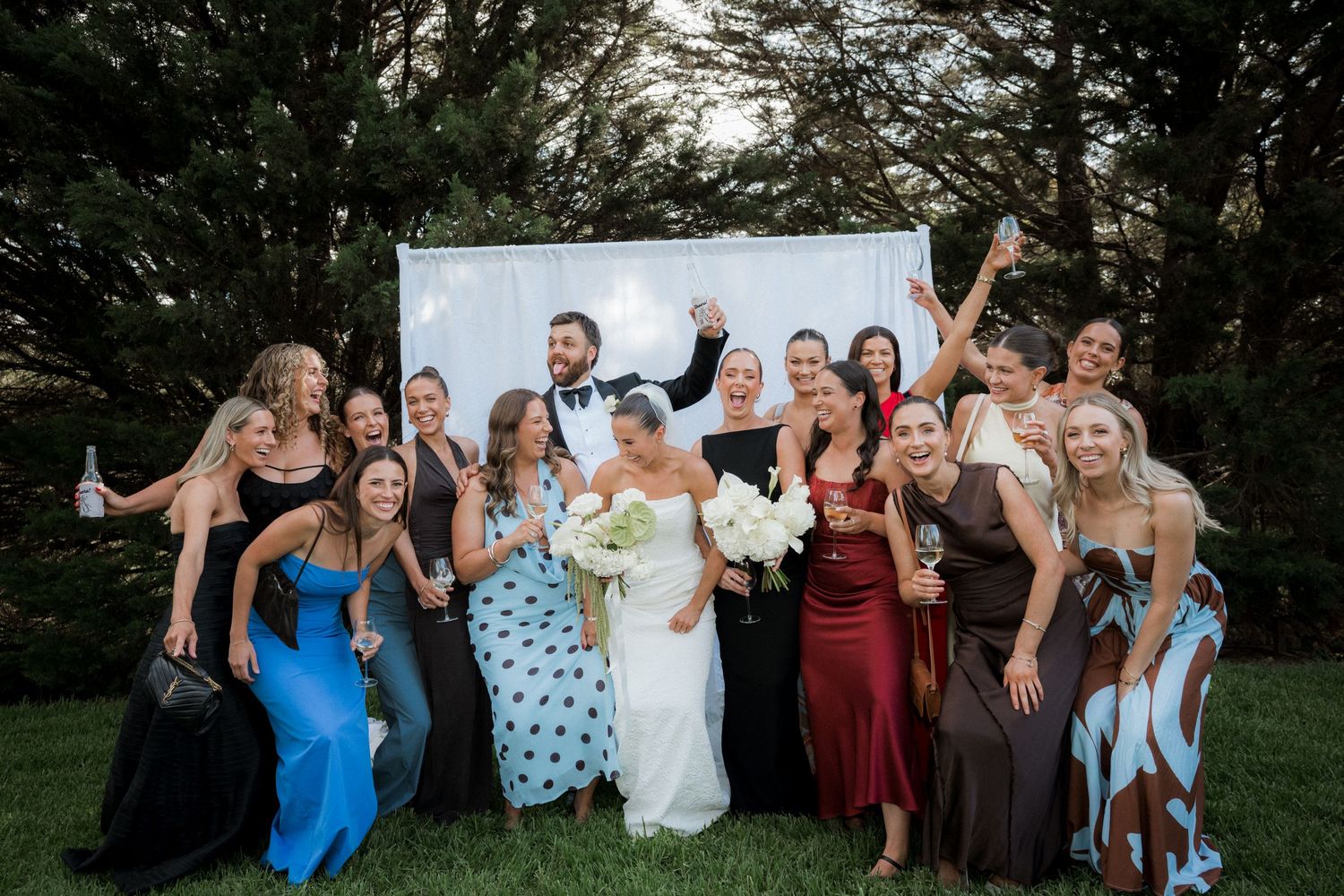 Bride and groom celebrate joyfully with wedding party guests in front of a white backdrop.