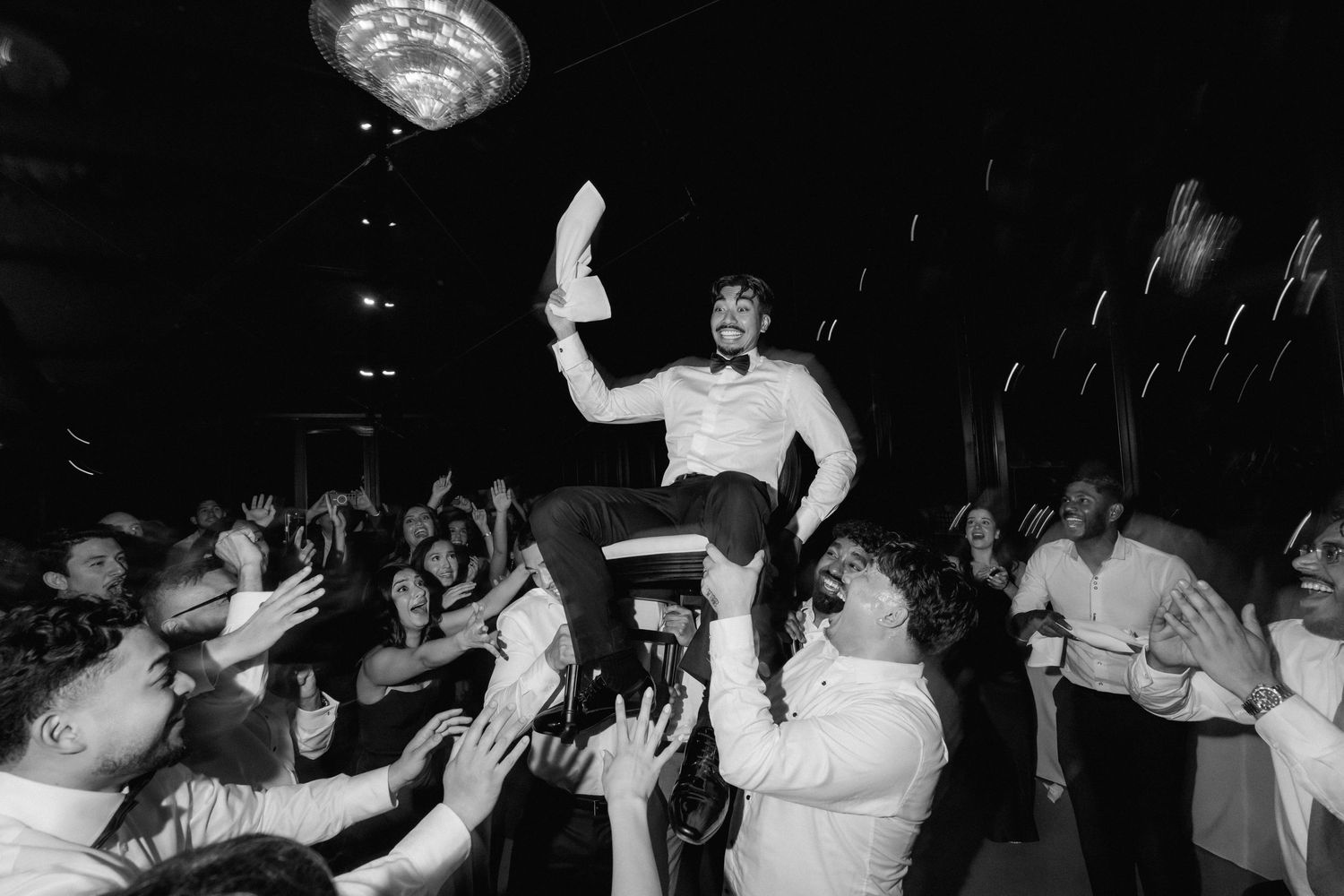 Black and white photo of a groom lifted on a chair by cheering wedding guests as he waves a napkin during a lively reception dance.