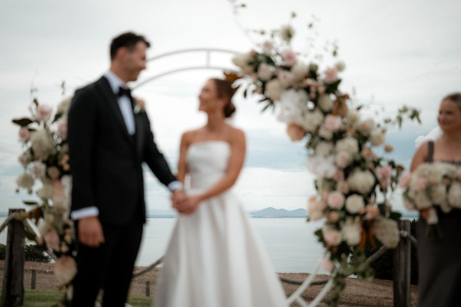 A blurred wedding ceremony takes place under a floral arch overlooking a waterfront view.