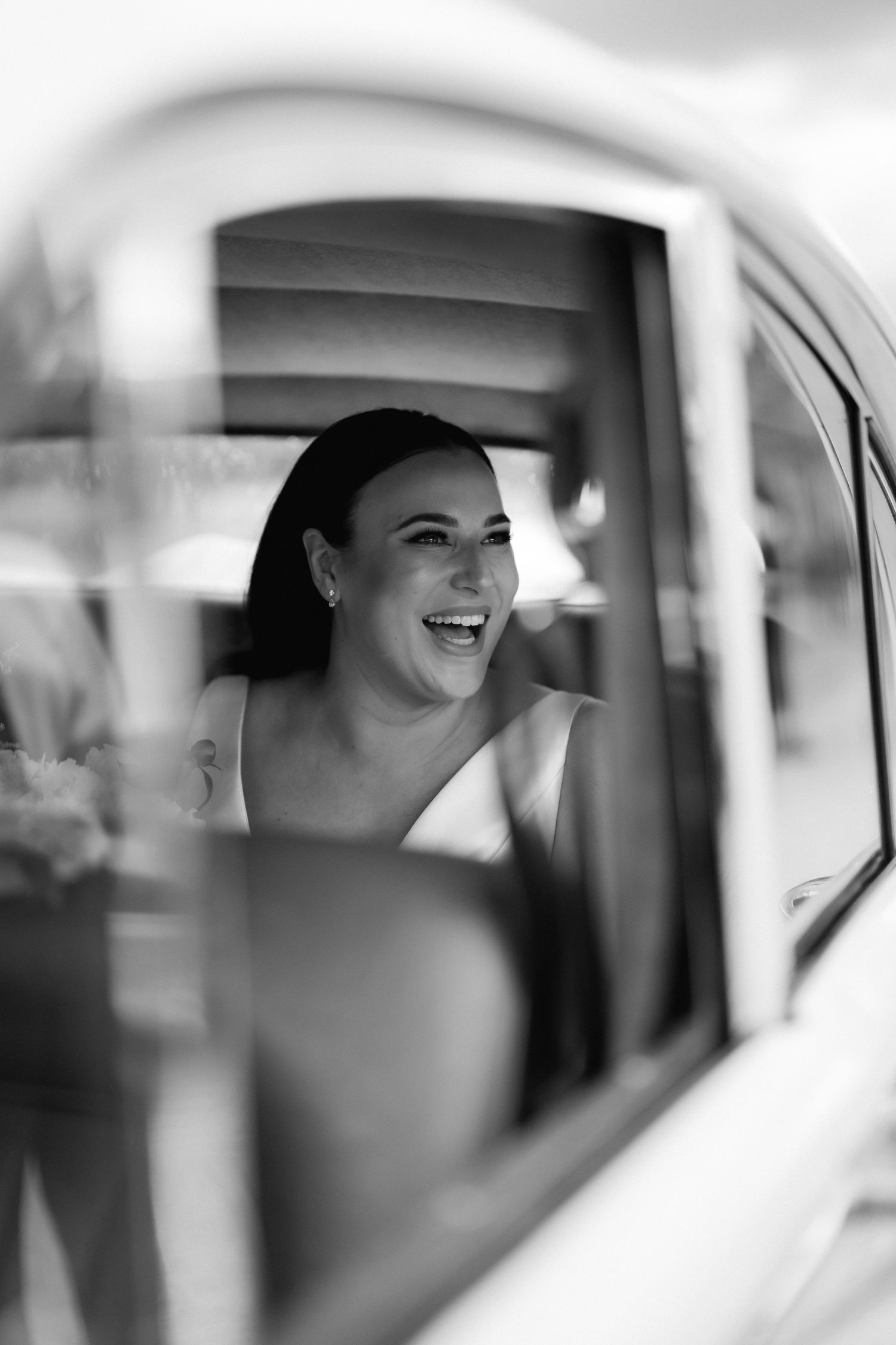Black and white portrait through a car window shows a woman in formalwear laughing joyfully.