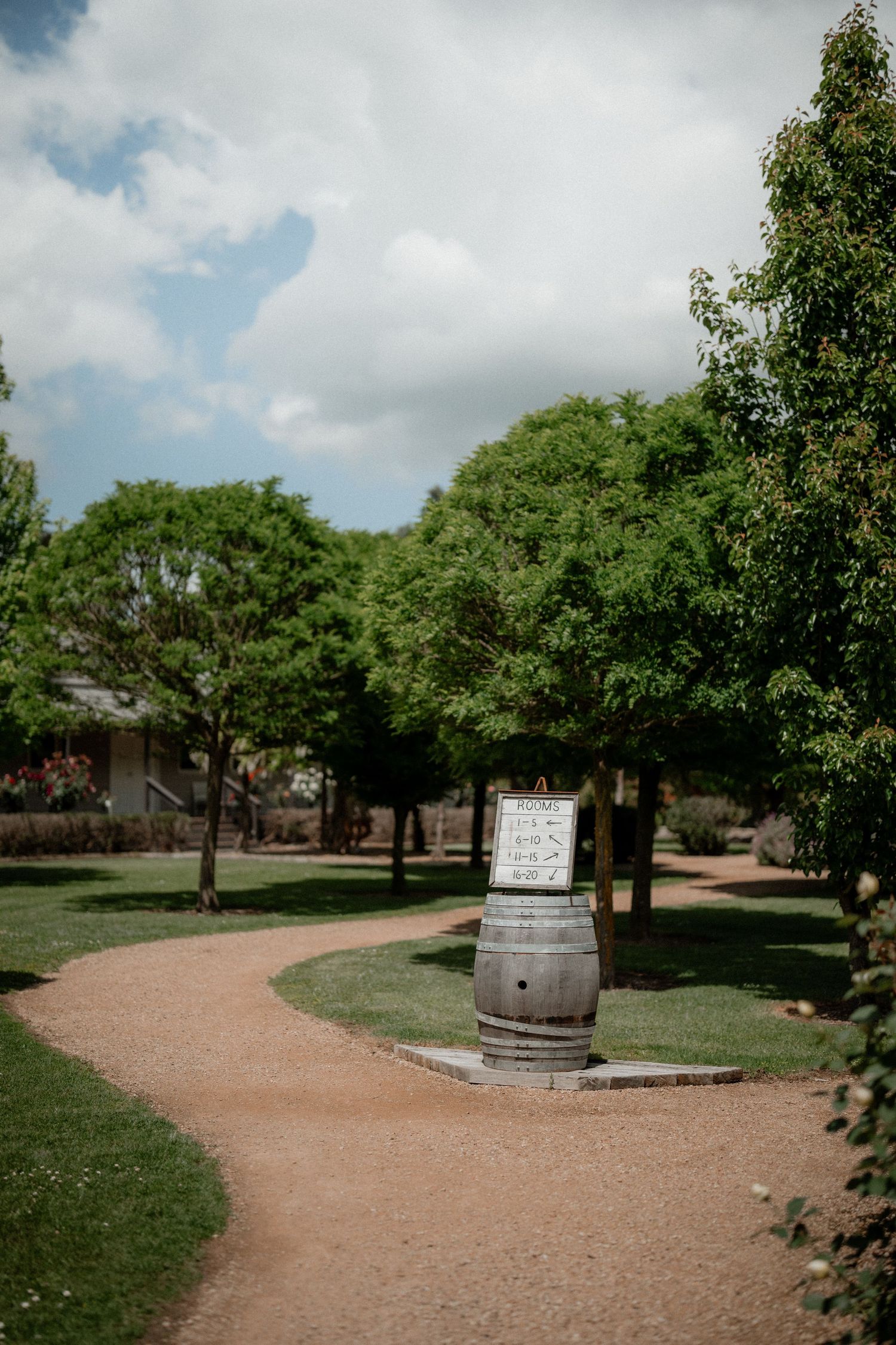 A winding dirt path leads through a green park with trees and a rustic wooden barrel display.