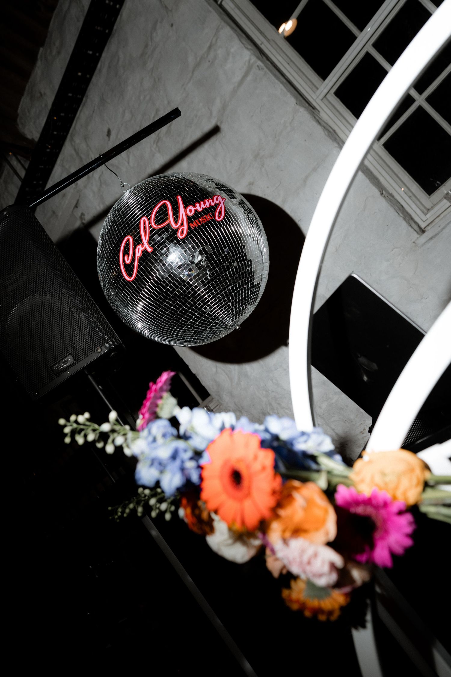 Disco ball with neon 'Get Young' text hangs above colorful floral arrangements at a party venue.