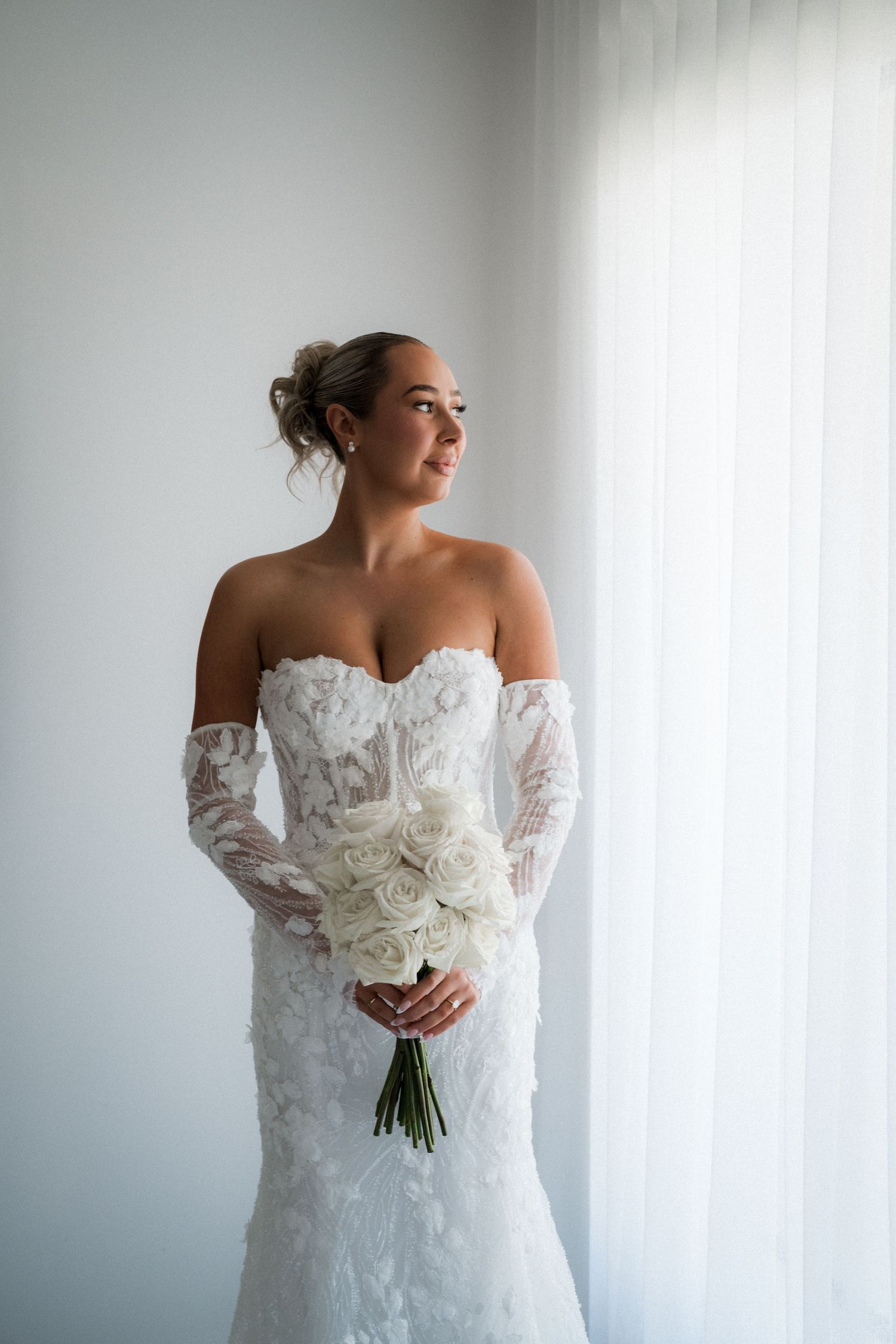 A bride in an elegant off-shoulder lace wedding gown holds a white rose bouquet against a light backdrop.