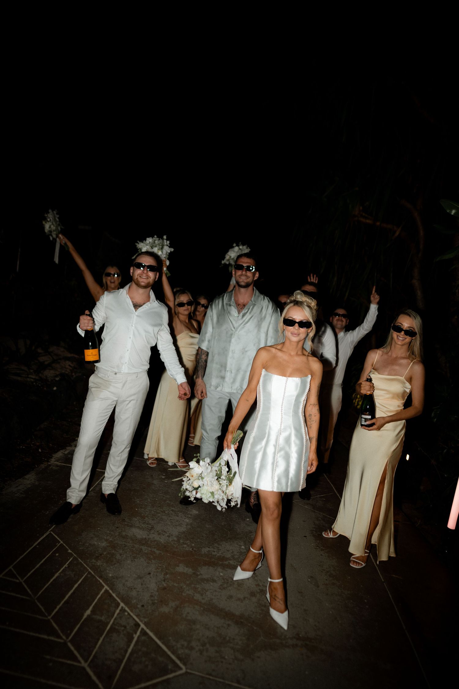 A wedding party in white and beige attire poses at night with champagne bottles and sunglasses.