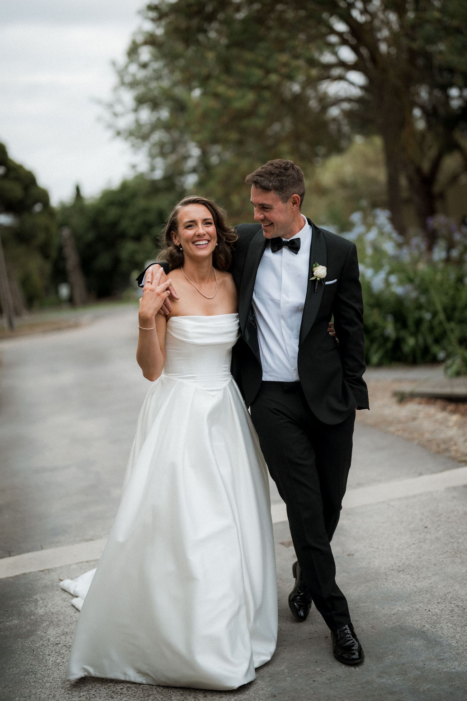 Newlyweds walk together and laugh on a tree-lined path.
