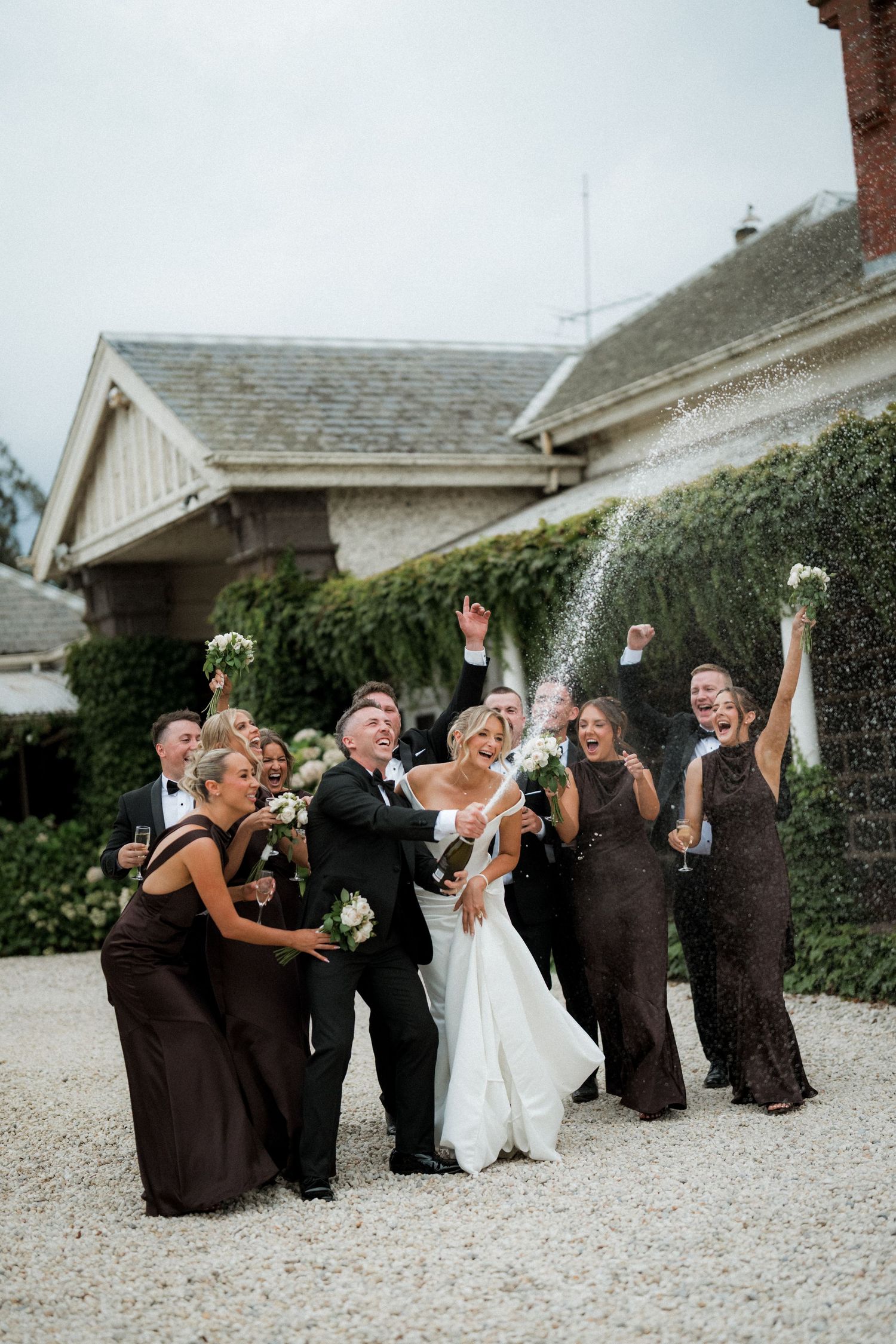 Wedding party celebrates with raised arms outside rustic venue.