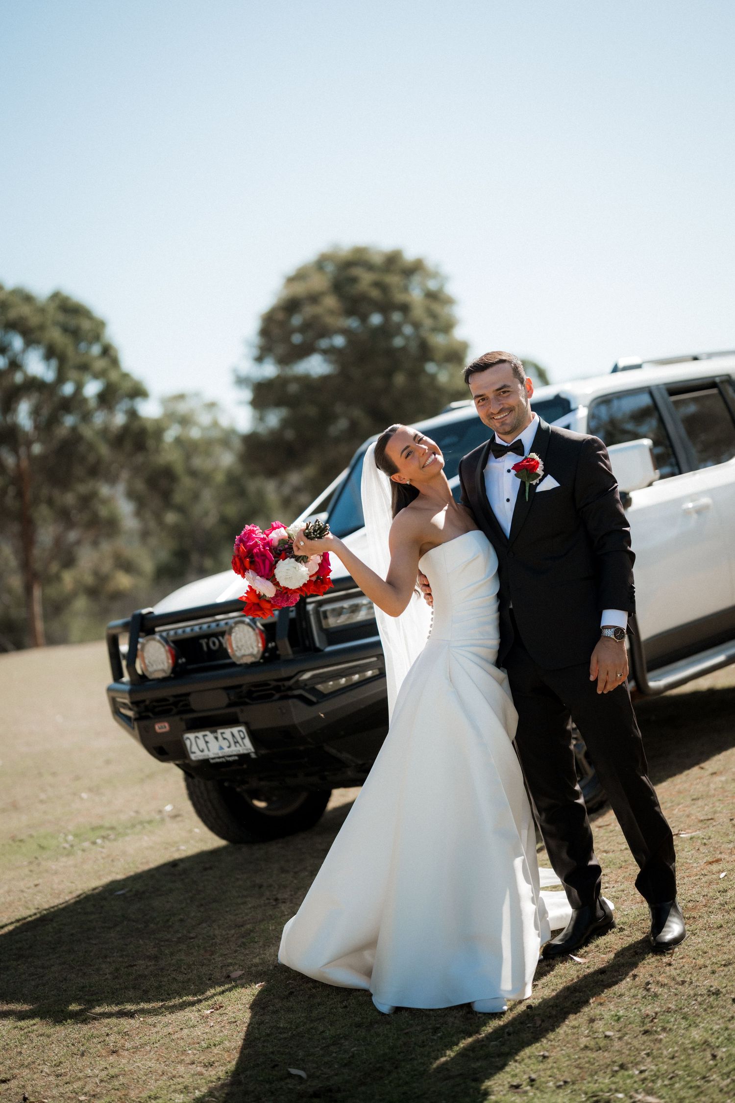 A bride and groom share playful moments near a black limousine in an outdoor wedding photoshoot.