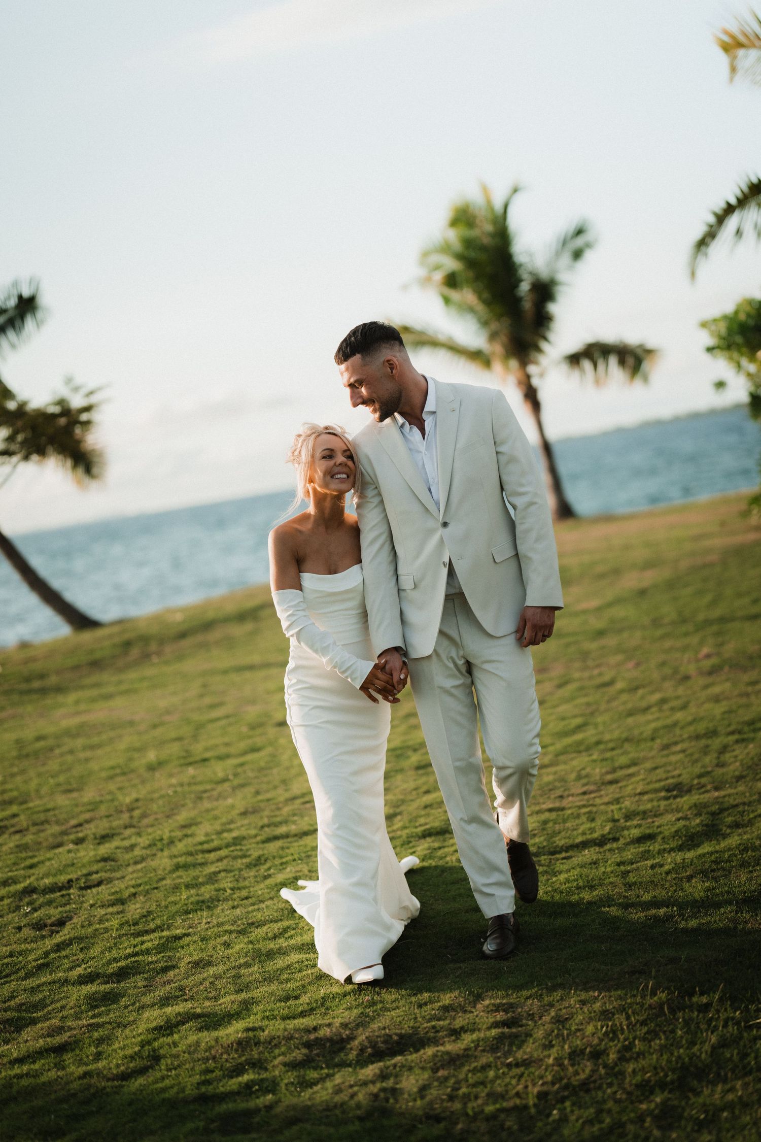 A couple in white attire walks along a grassy hillside with palm trees and ocean views at sunset.