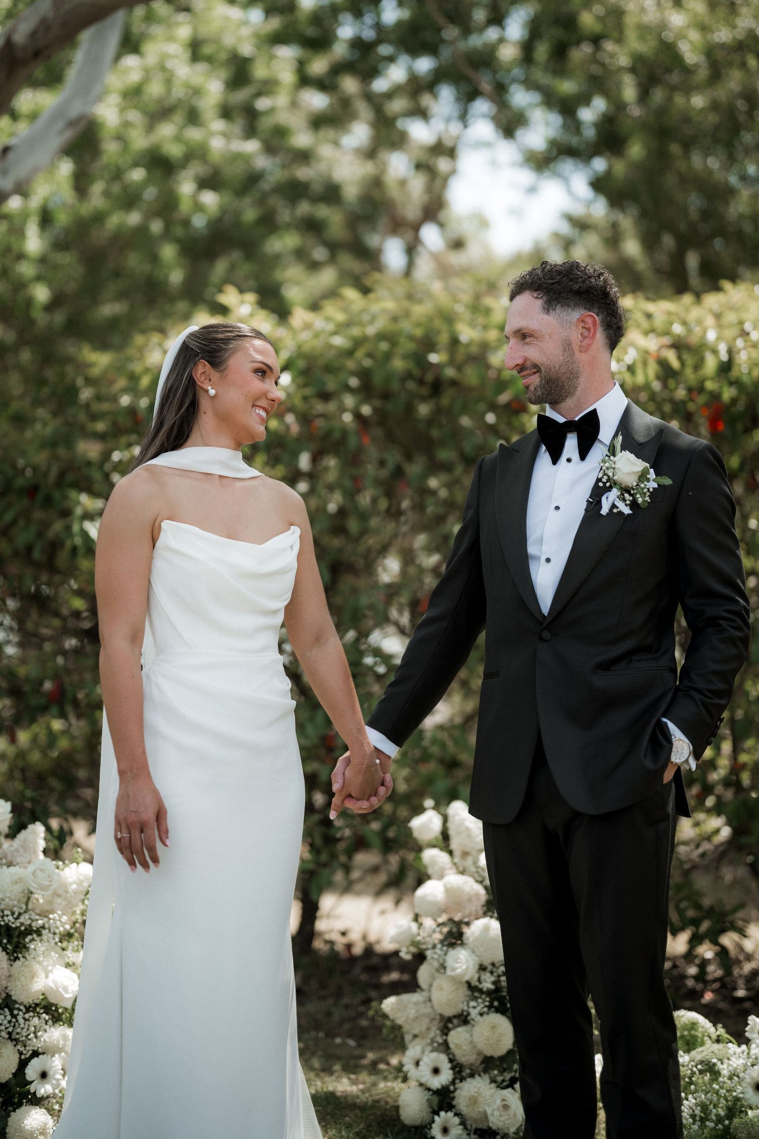 Elegant wedding couple poses together outdoors in a garden setting with white floral arrangements.