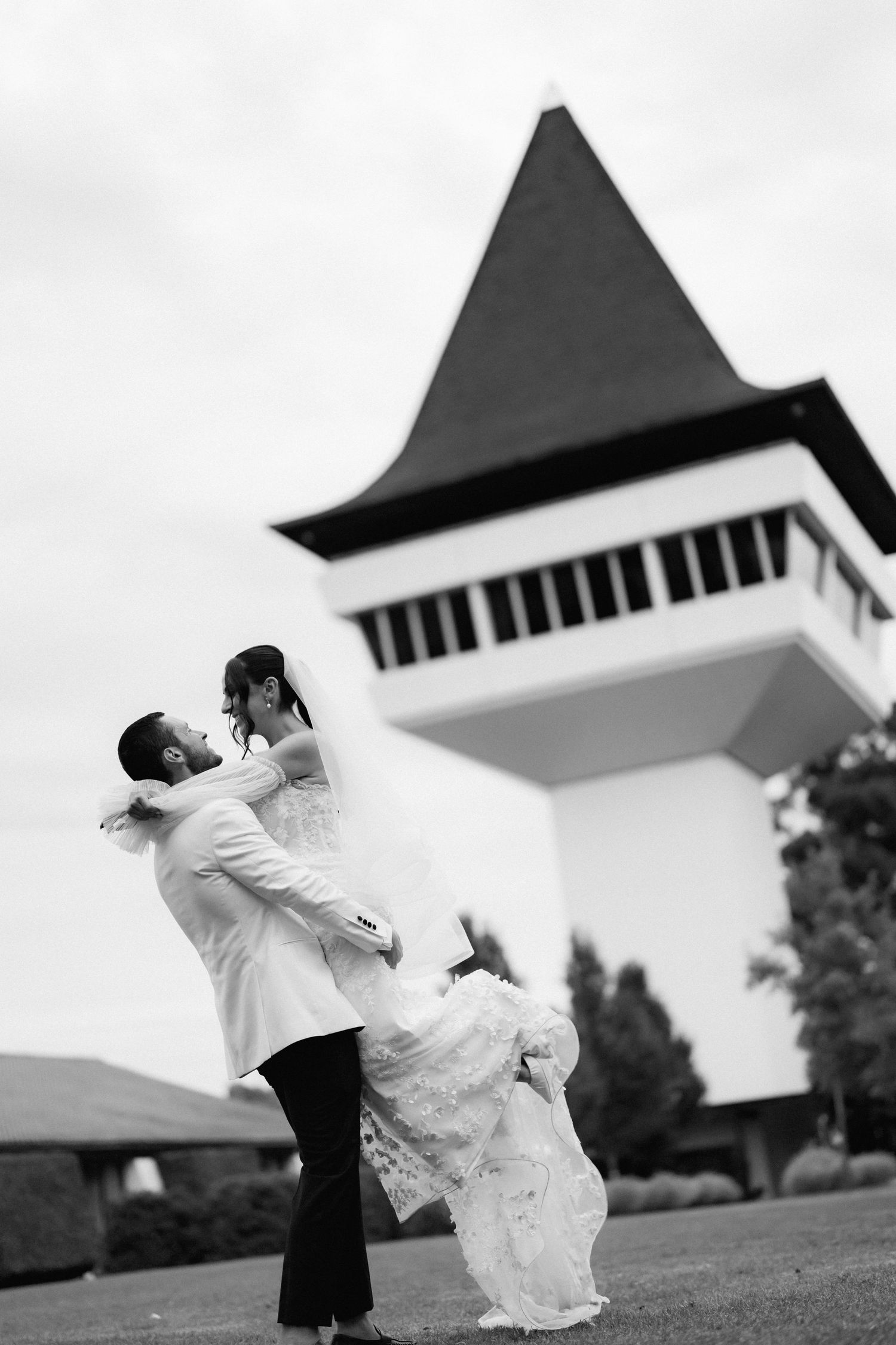 Newlyweds share a romantic dance moment on an outdoor lawn with a distinctive peaked roof building behind them.