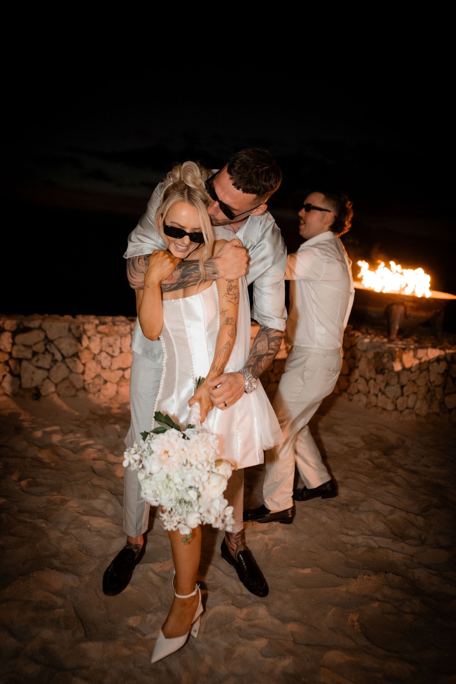 Wedding celebration at night with couples dancing near a fire pit on sandy beach.
