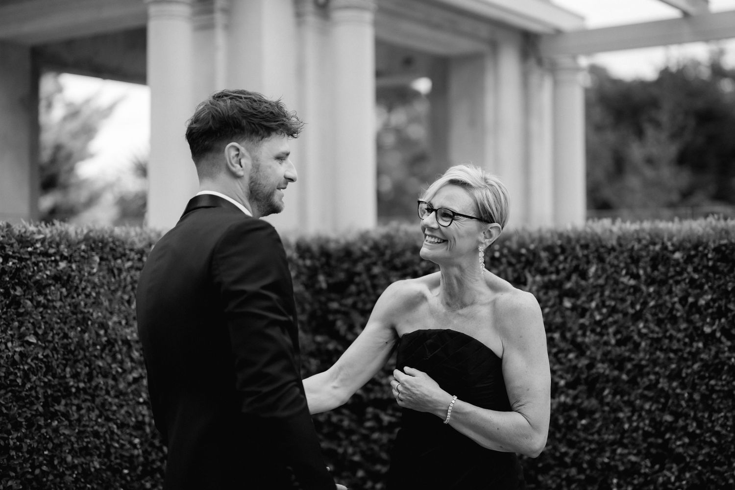 Two people share an intimate moment during a formal event in front of elegant columns and manicured hedges.