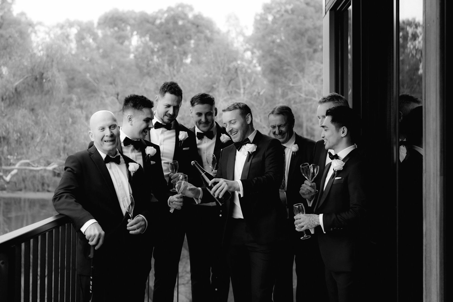 Group of groomsmen in black tuxedos gather on a balcony for candid moment during wedding celebration.