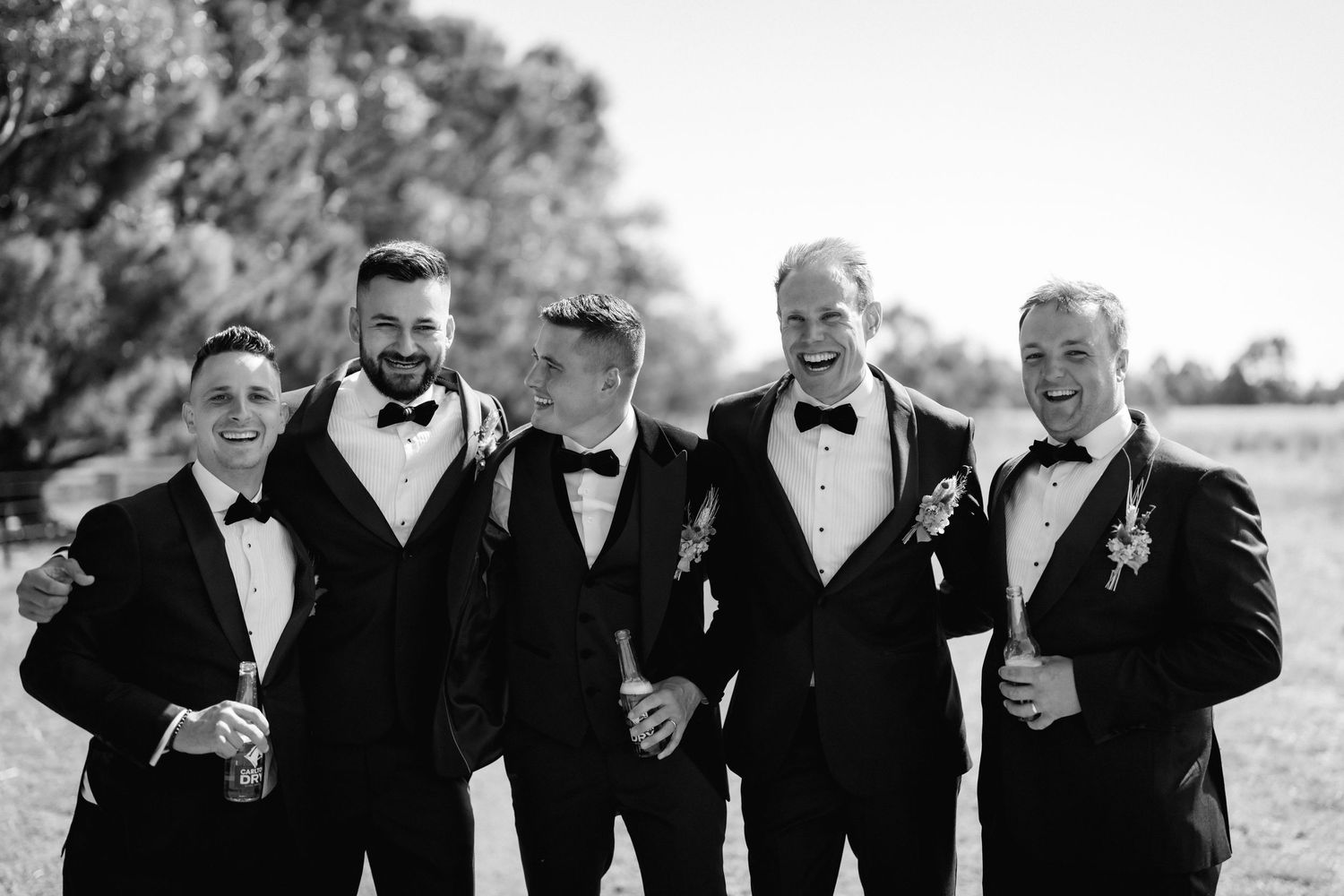 A black and white photo of five groomsmen in tuxedos laughing together at an outdoor wedding.