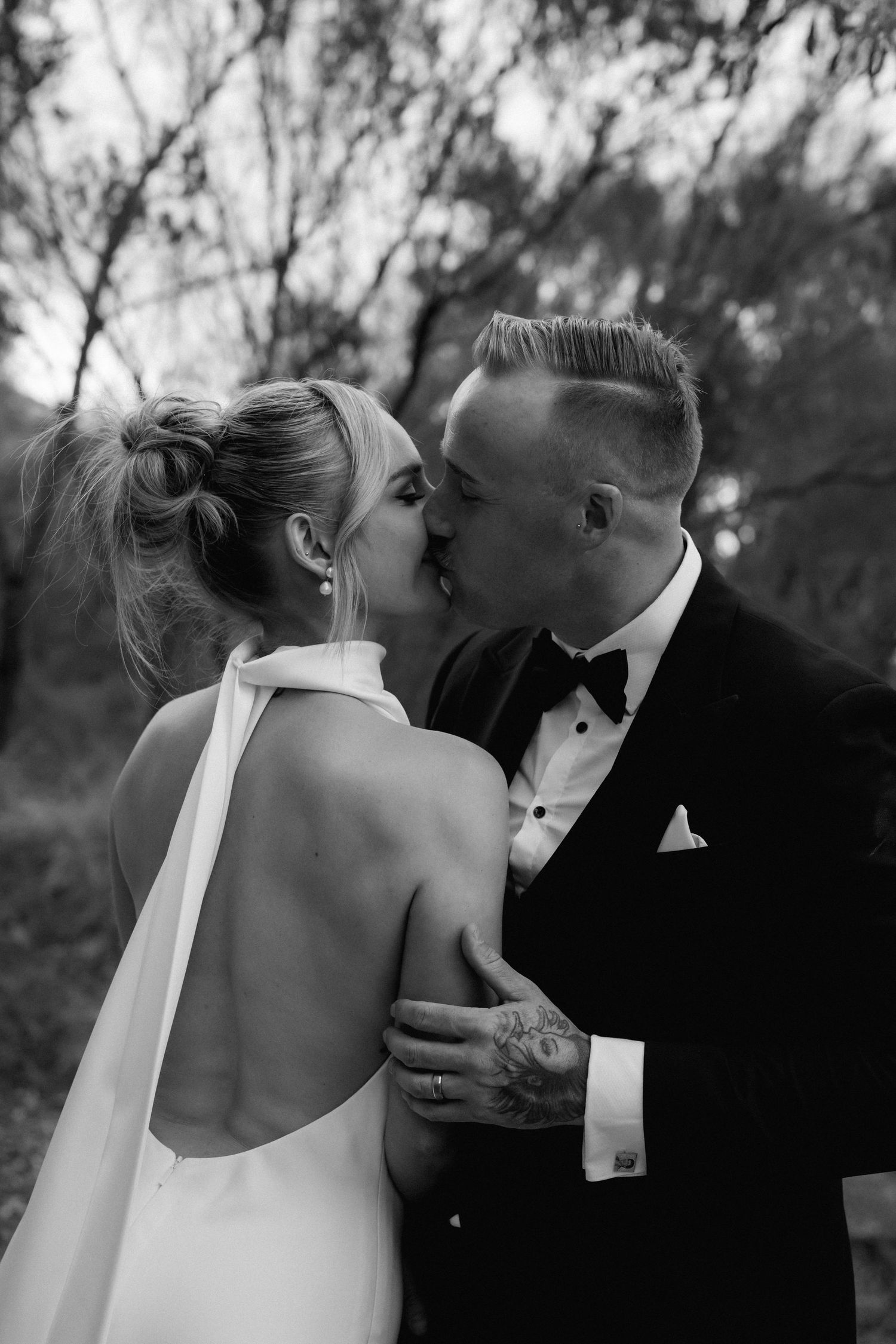 Black and white romantic portrait of newlyweds kissing, bride in backless dress and groom in classic tuxedo.