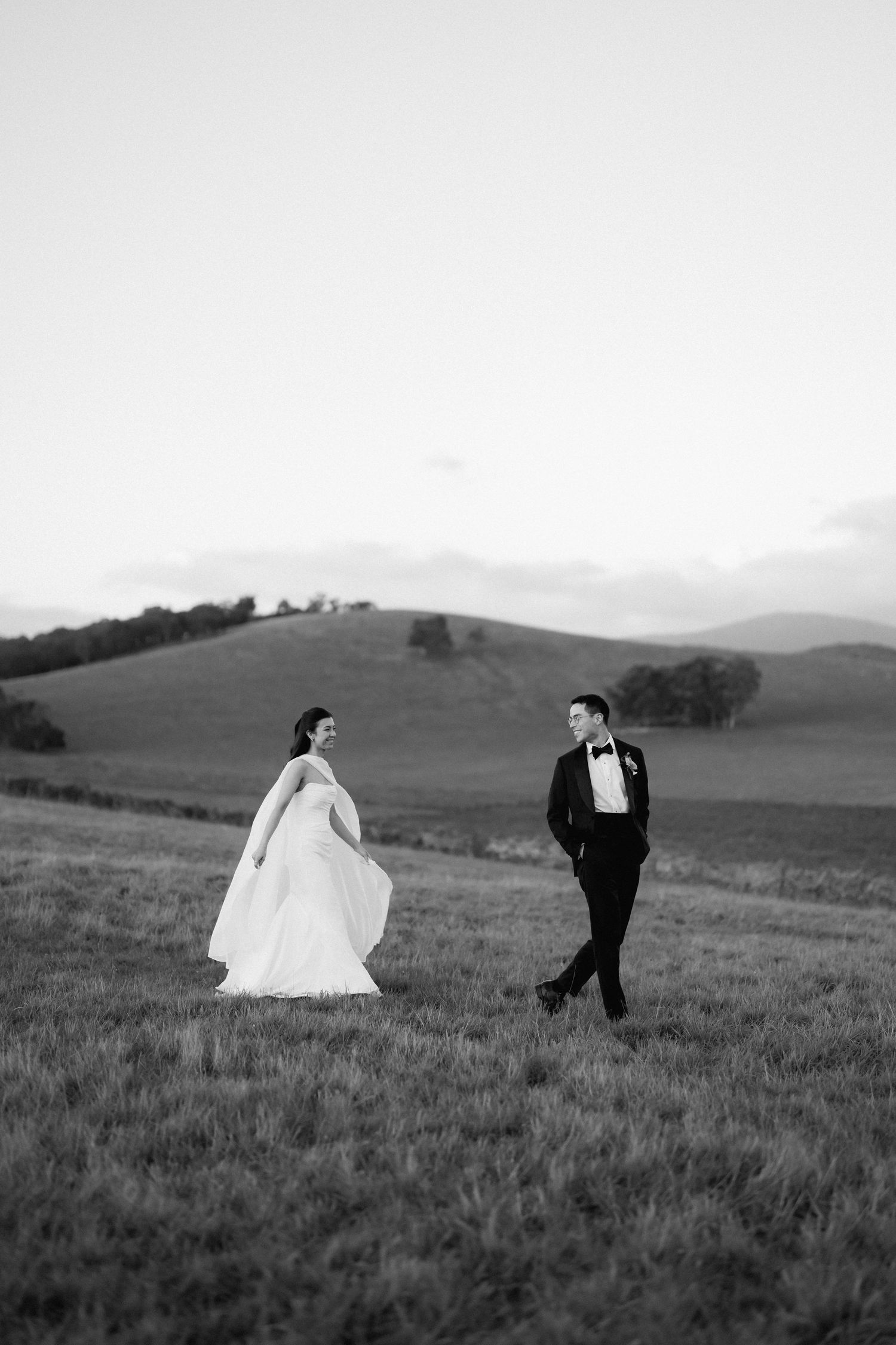 Black and white photo of bride and groom walking through open countryside fields with rolling hills at dusk.