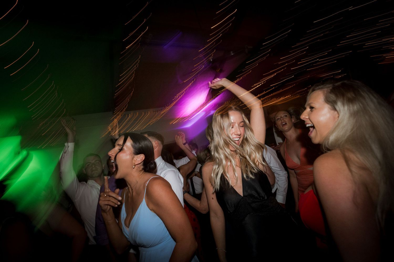 Party guests dancing under colorful purple and green lighting at a nighttime celebration.
