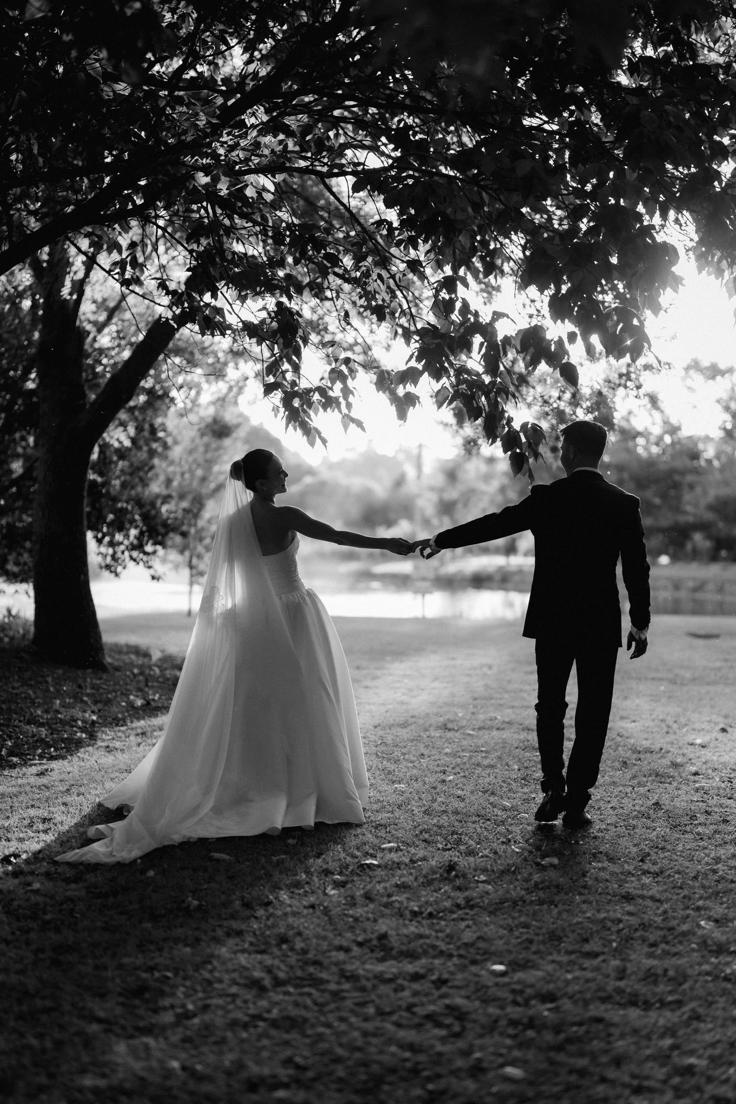 Silhouetted couple in wedding attire walking under trees at sunset.