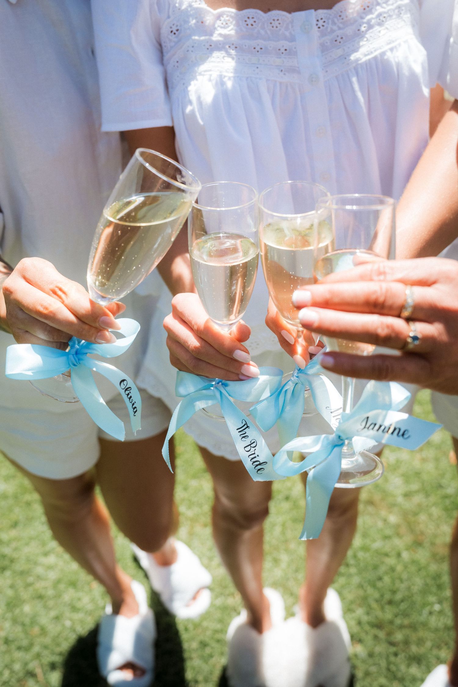Row of champagne glasses with blue ribbons held by bridesmaids in white dresses on grass.