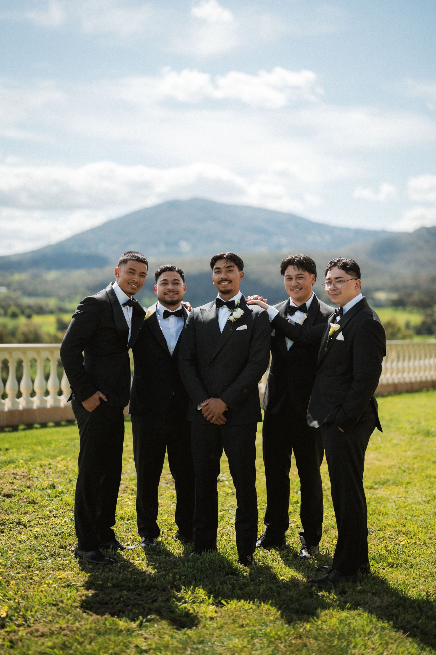 Five groomsmen in black tuxedos and bow ties pose together on a sunny lawn with a mountain and cloudy sky behind them.