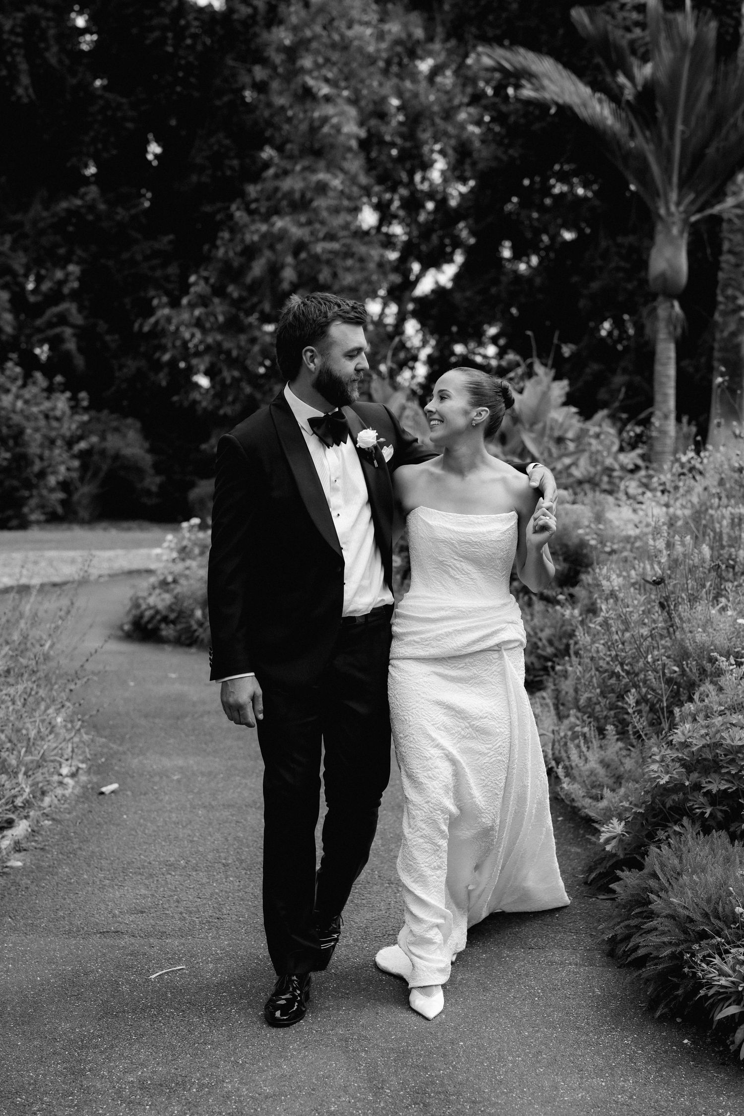 Black and white photo of bride and groom walking arm in arm along a garden pathway on wedding day.