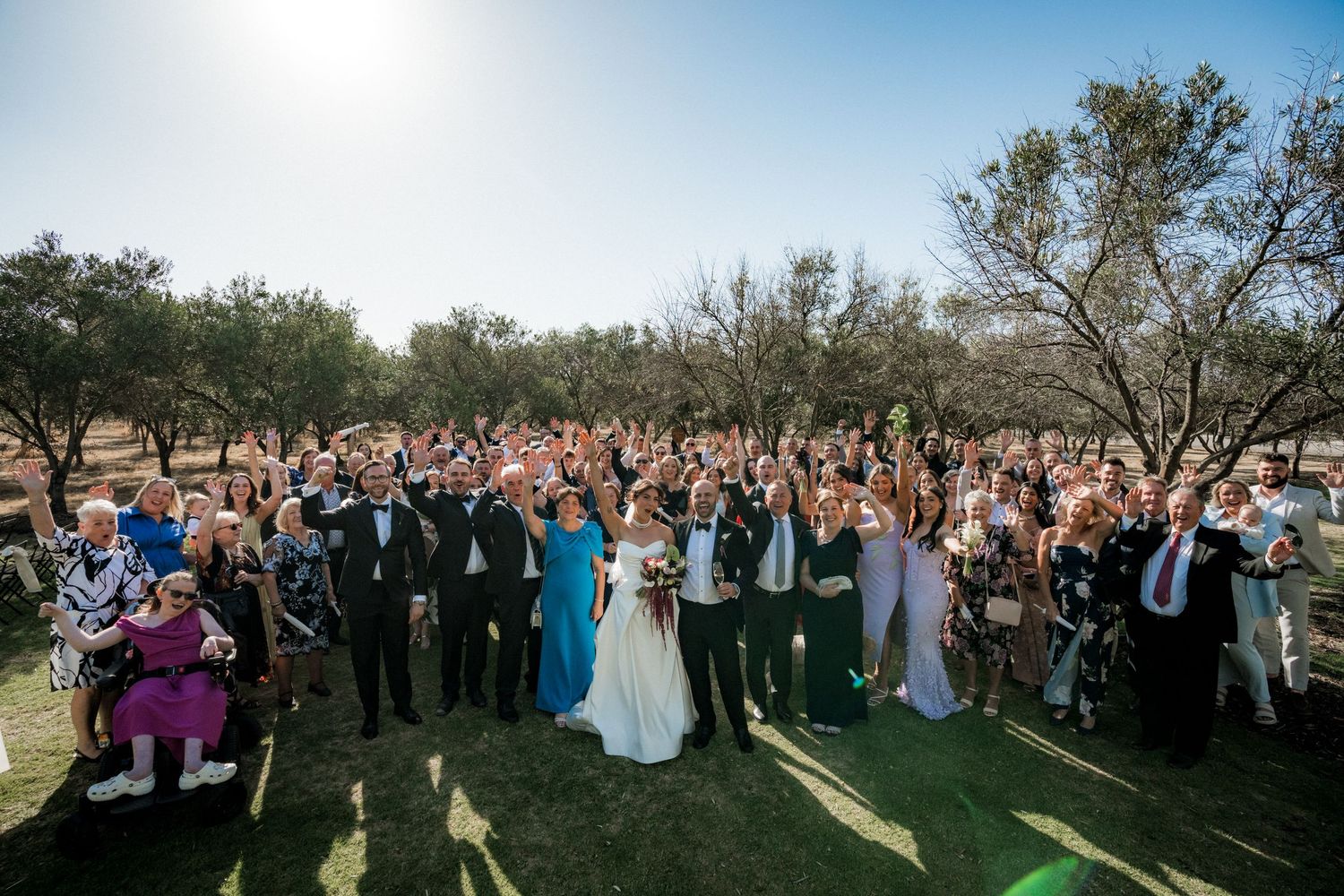 Wedding guests gather outdoors in a scenic natural setting with trees and sunlight filtering through the branches.