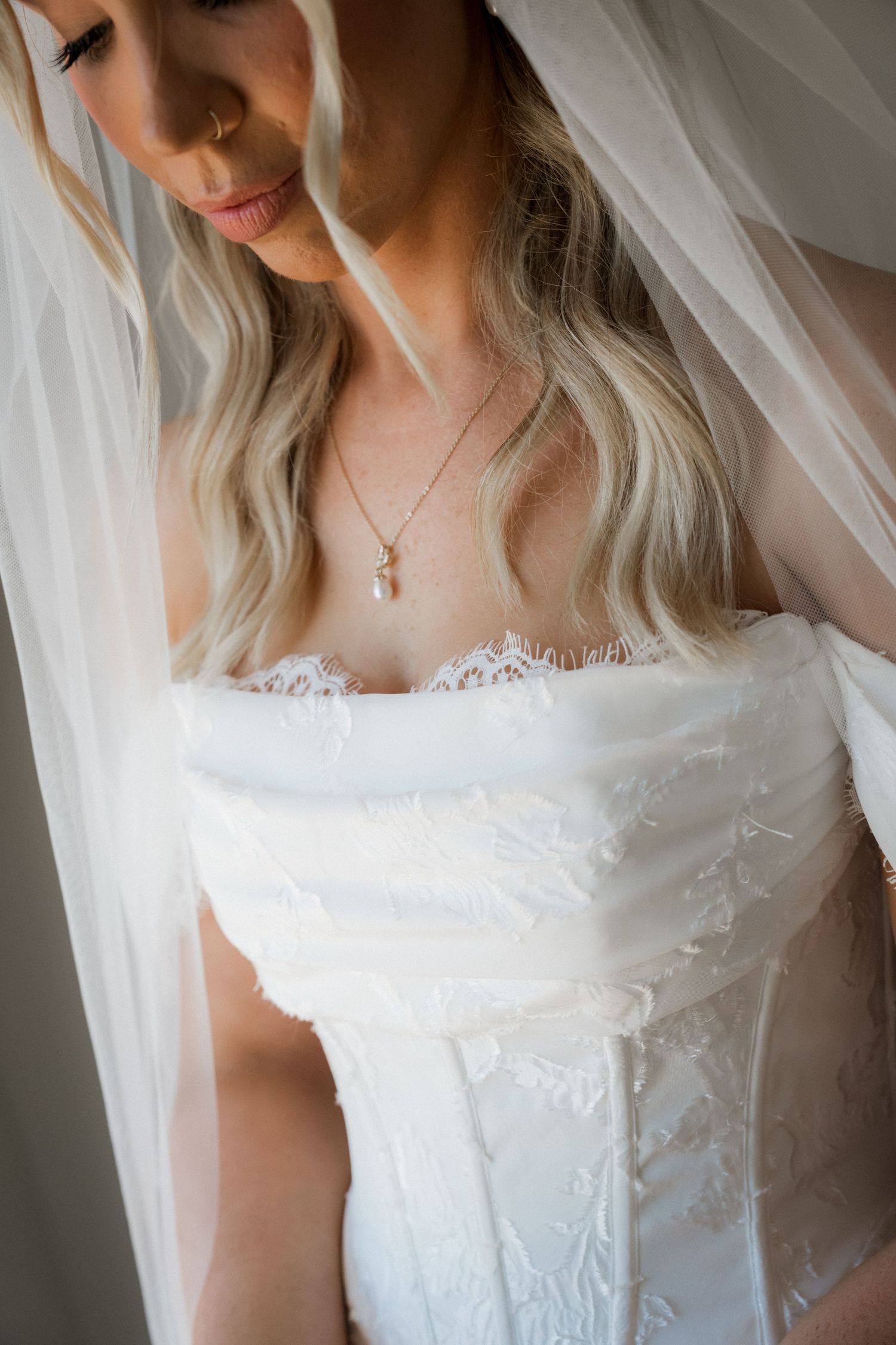 Bride in strapless lace wedding dress and veil with gold necklace looking down elegantly.