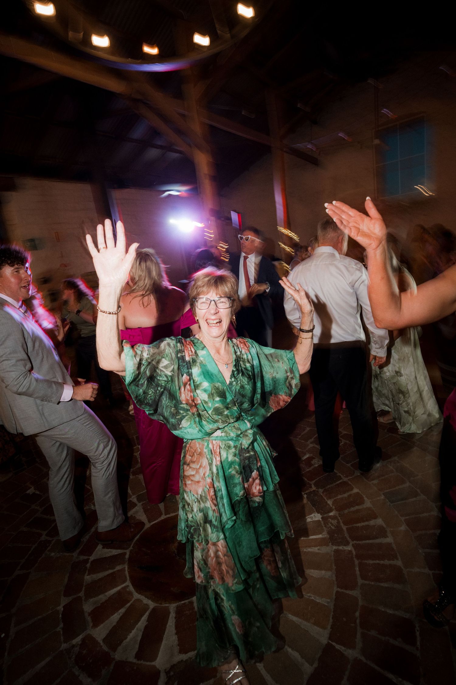 Elderly woman in green floral dress dancing joyfully at wedding reception with hands raised.