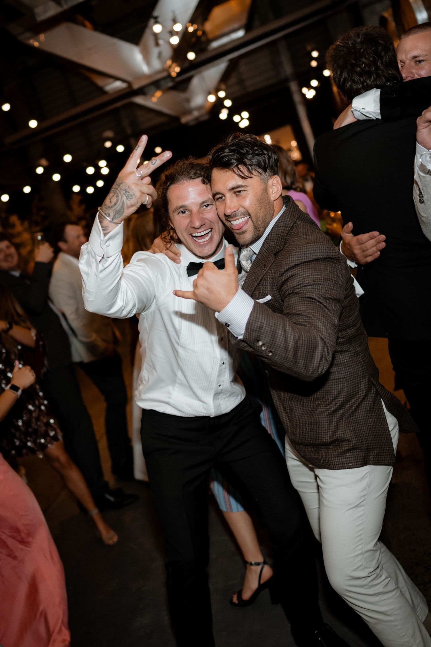 Two wedding guests celebrating and dancing together at reception with string lights overhead.