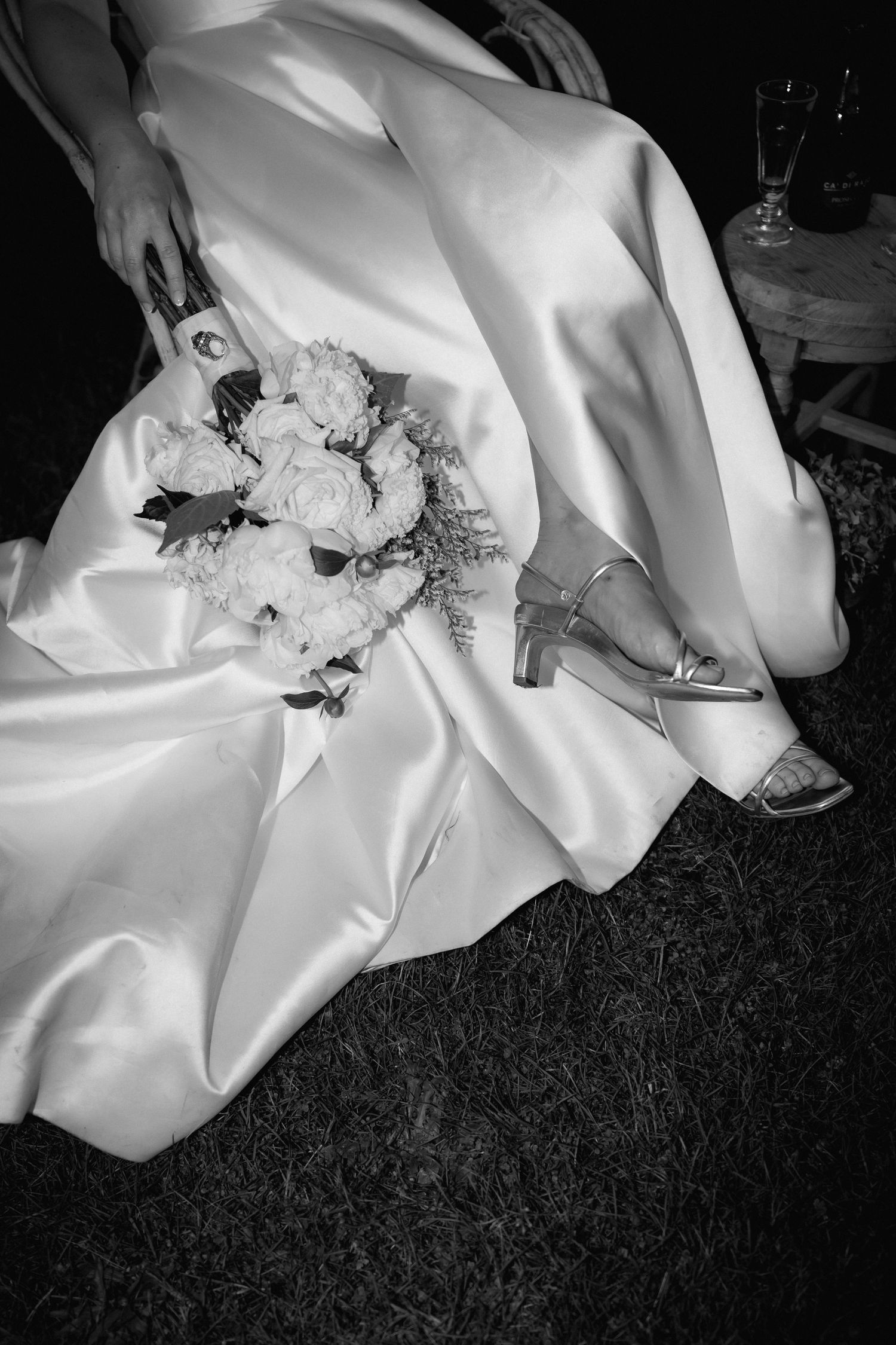 A bride's elegant white wedding dress flows over grass while holding a bouquet of white flowers and wearing silver shoes.