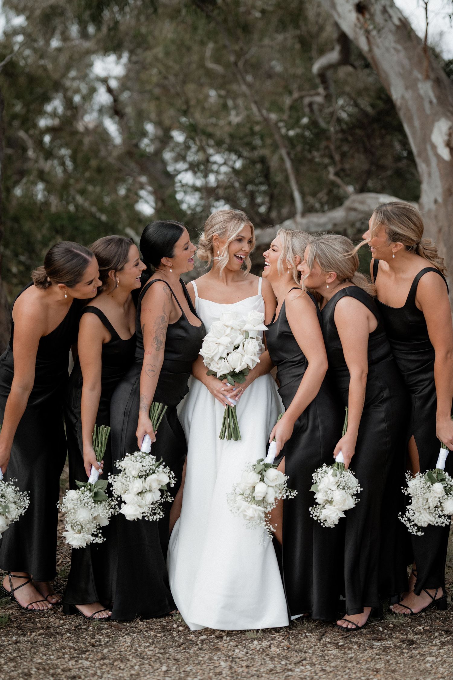 Bridal party in black dresses holding white bouquets stand in a line against a rustic outdoor background.