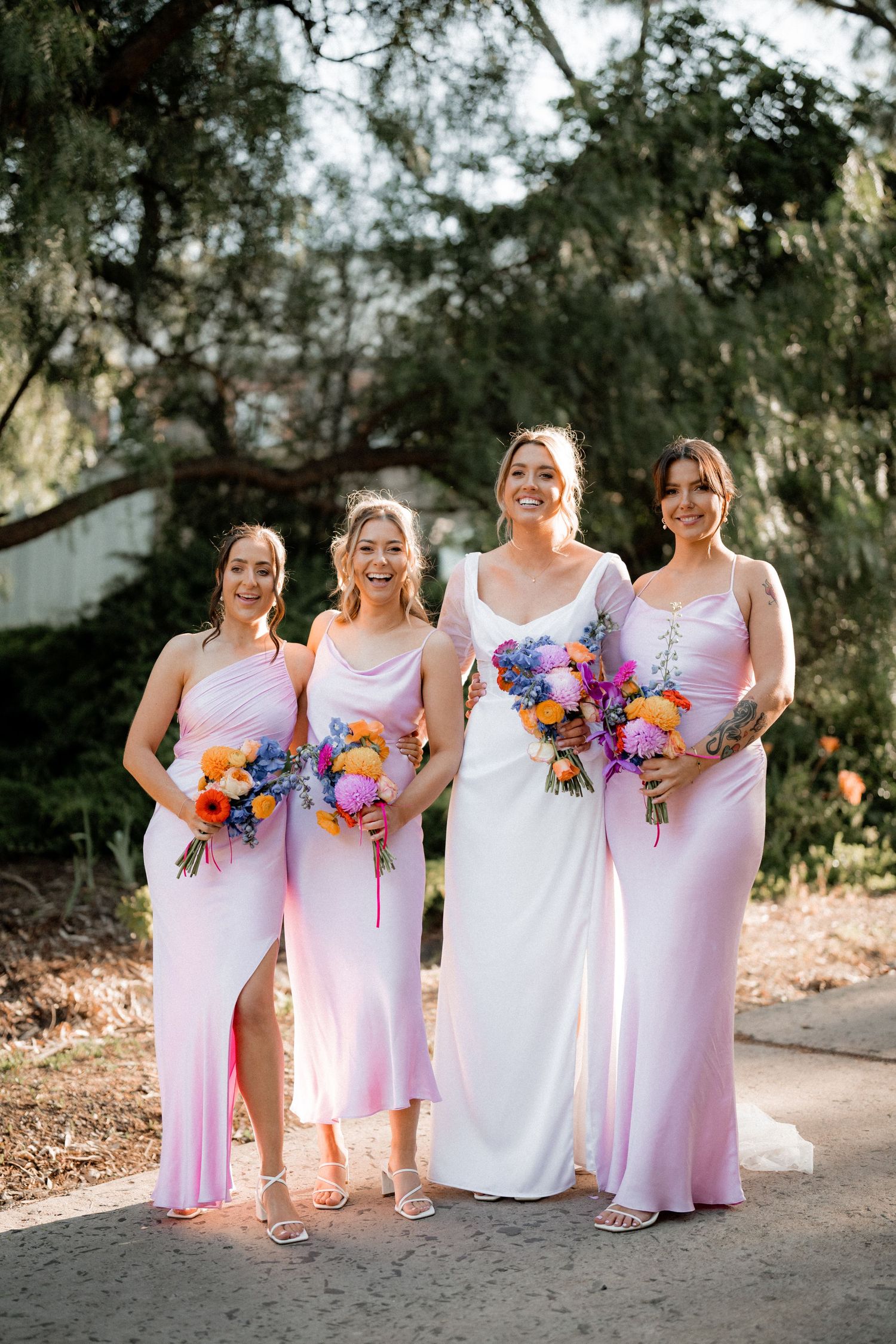 Wedding party wearing pink dresses and holding vibrant floral bouquets pose together on a sunlit outdoor path.