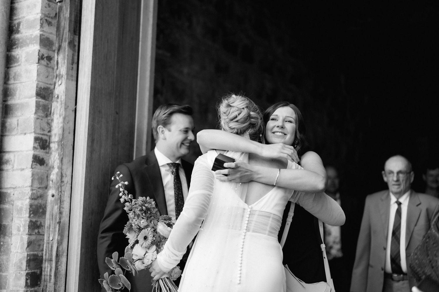 A black and white candid moment captured during a wedding celebration outside a brick building.