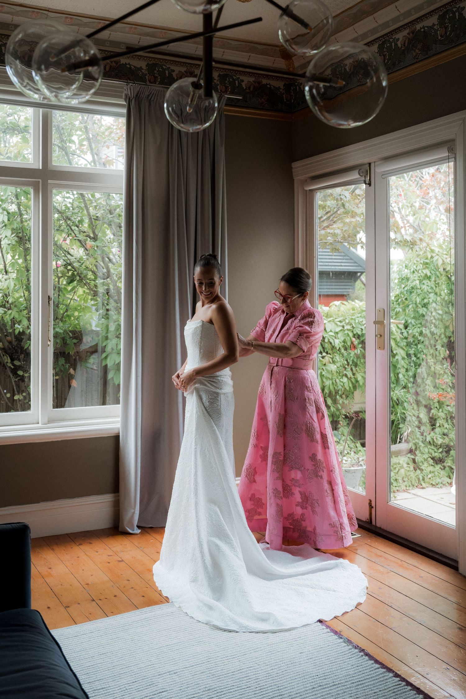 Bride in white gown being helped by woman in pink floral dress near large windows in elegant room.