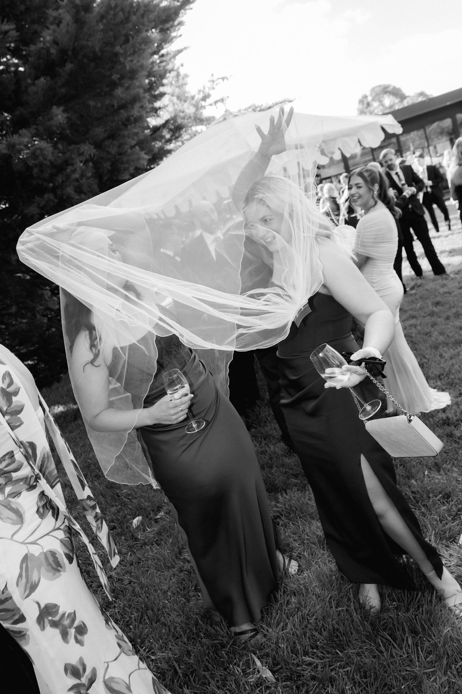 Bride laughing under veil with bridesmaids in black dresses at outdoor wedding reception in black and white photo.