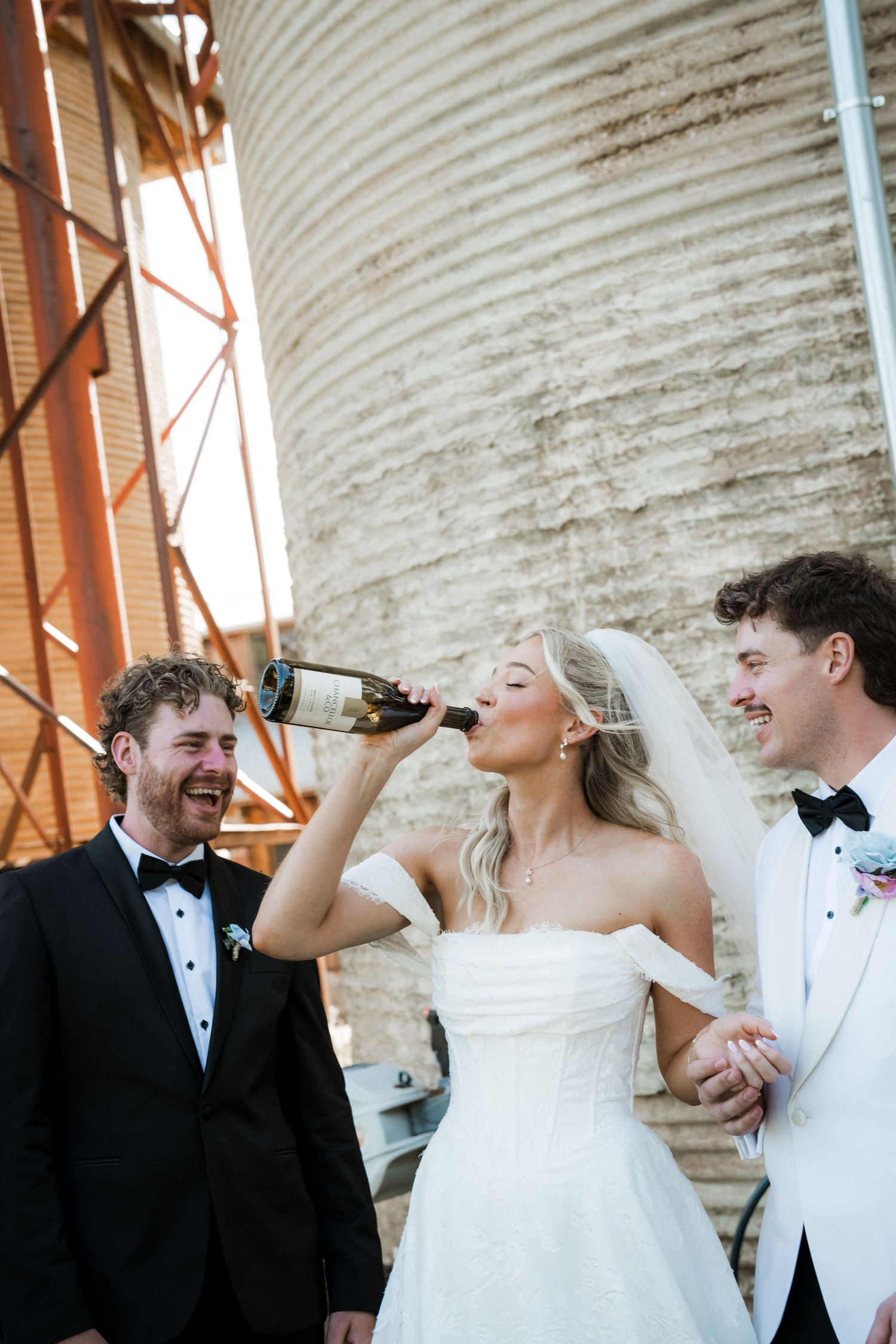 Bride drinks champagne bottle while groom and groomsman laugh beside industrial grain silo at wedding.