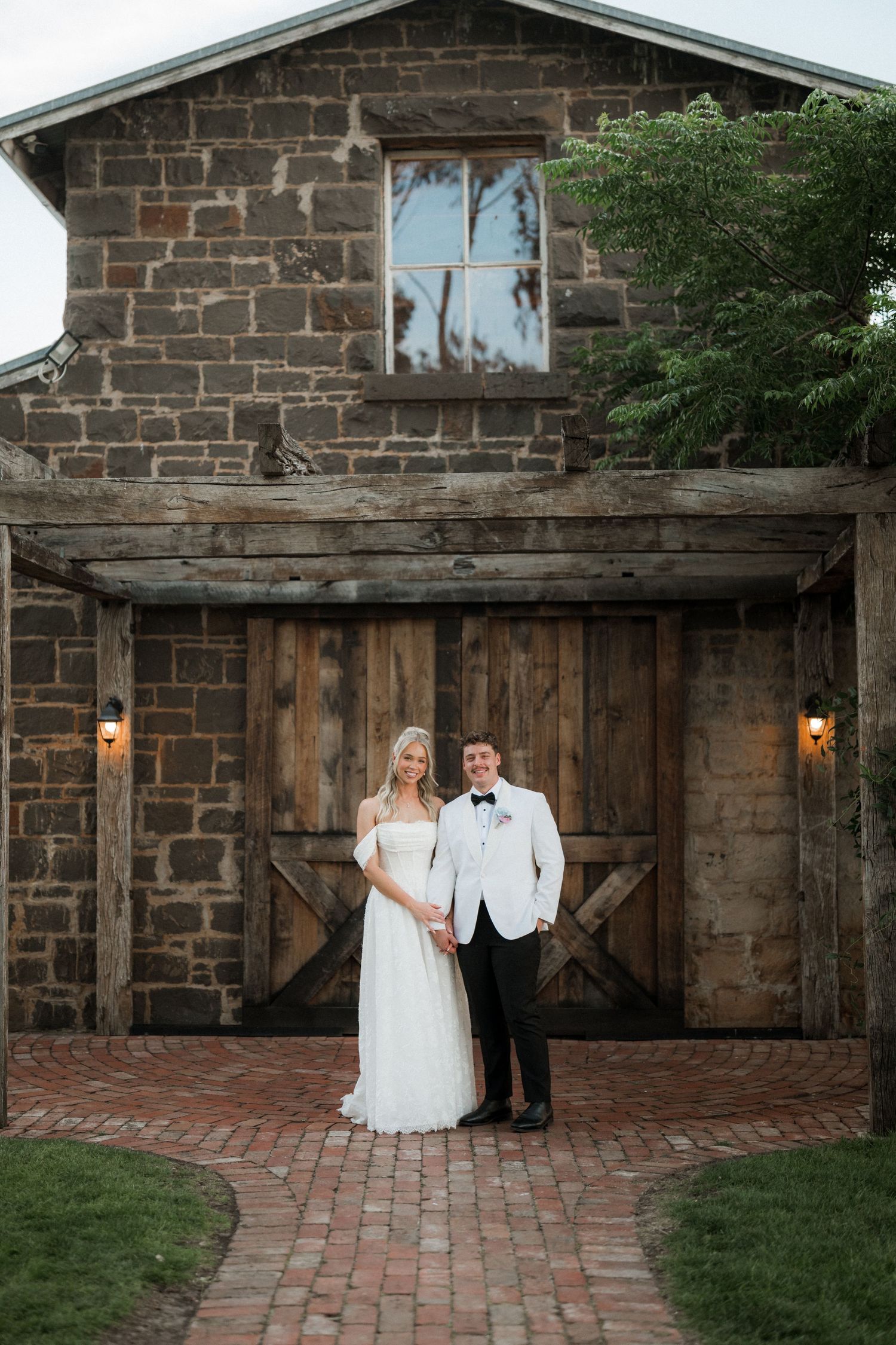 Bride and groom pose hand in hand before rustic stone barn with large wooden doors at wedding.