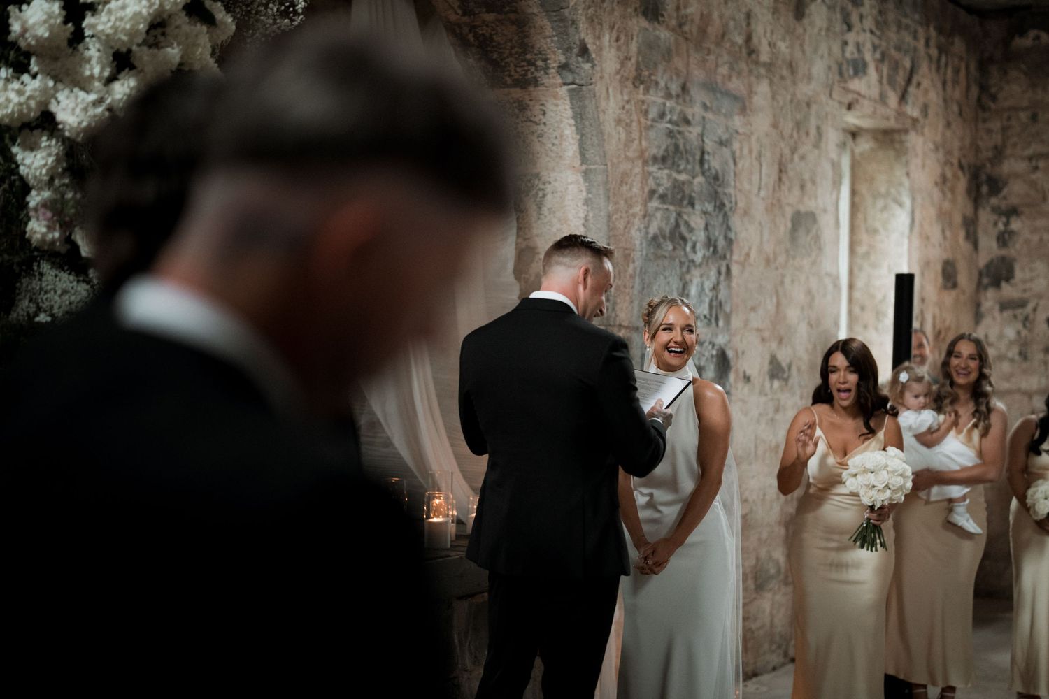 A couple exchanges vows during an intimate wedding ceremony surrounded by bridesmaids in a stone-walled venue.