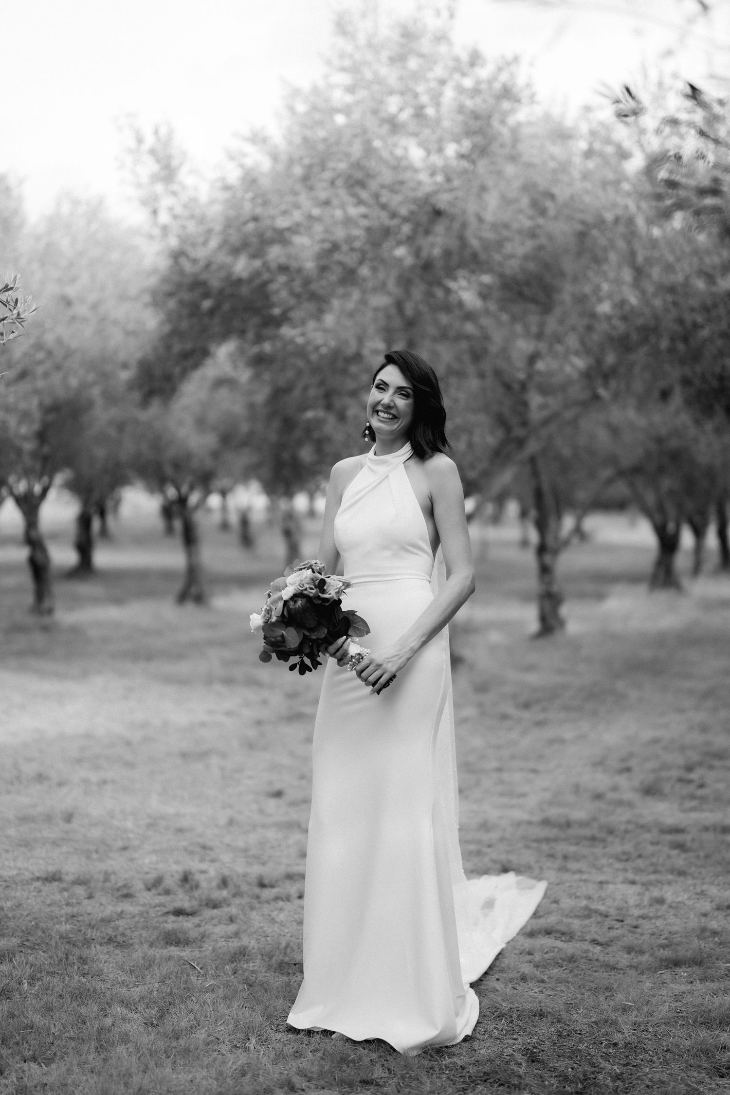 Black and white portrait of a bride holding bouquet while standing in an orchard setting.